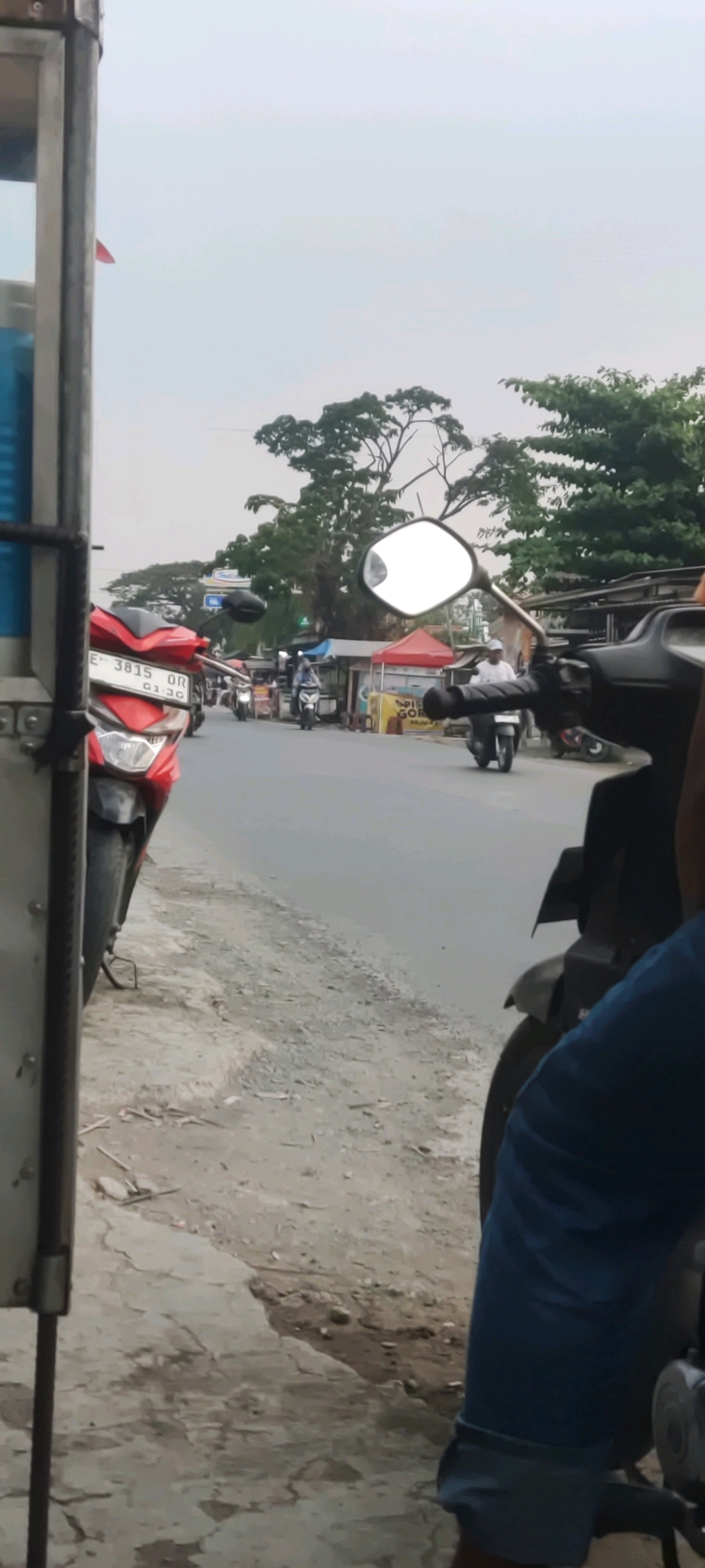 The image depicts a street scene in Gebang, Indonesia. In the foreground, to the left, a portion of a metal cart or stand is visible. To the right of the cart, a red motorcycle is parked. The motorcycle's license plate reads "E 3815 OR".
Further down the paved road, several other motorcycles are in motion. One is being ridden by a person wearing a blue shirt. Another motorcycle, closer to the right edge of the image, is partially obscured by the viewpoint. On the right side of the road, there are small stalls with red and white awnings. Trees and buildings line the background. The sky is overcast.