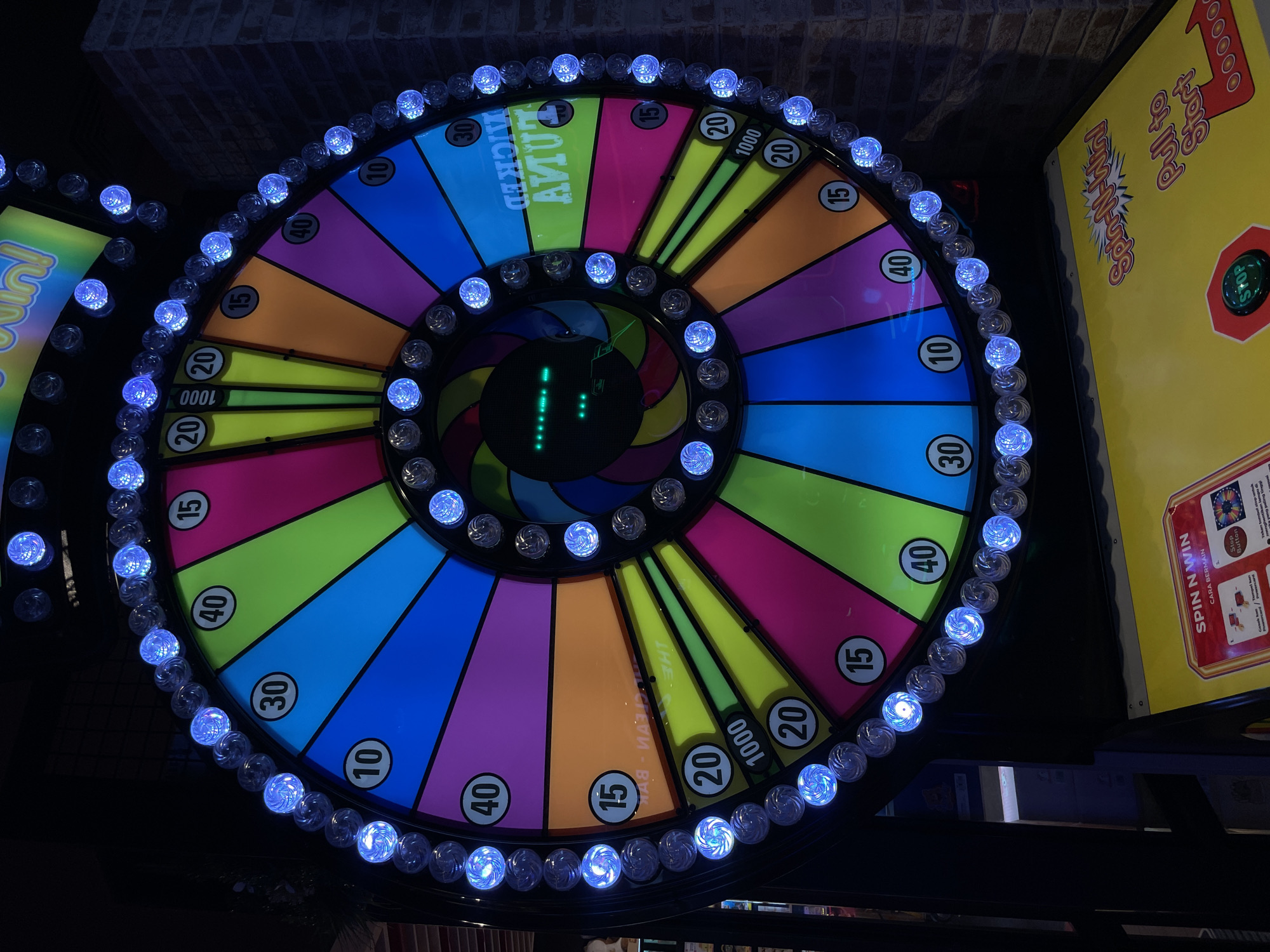 The image shows a brightly lit carnival-style spinning wheel game, likely a "Spin N' Win" or similar, set up for play. The wheel is divided into numerous colorful segments, each featuring a number or prize indication such as 10, 15, 20, 30, 40, and 1000. The wheel is outlined with a string of blue LED lights that are illuminated, creating a festive and attention-grabbing display. In the center of the wheel, a digital display shows a series of green lights. At the bottom of the image, the control panel for the game is visible, featuring a yellow surface with the text "Spin N' Win" and instructions, along with a red button labeled "Stop." The game is set against a dark background, with brickwork visible on the right side of the image, suggesting an indoor entertainment venue or arcade. The overall scene depicts a recreational activity. The location is identified as Manado, Indonesia, likely within an entertainment establishment in the city. There are no people directly interacting with the game in the image.
