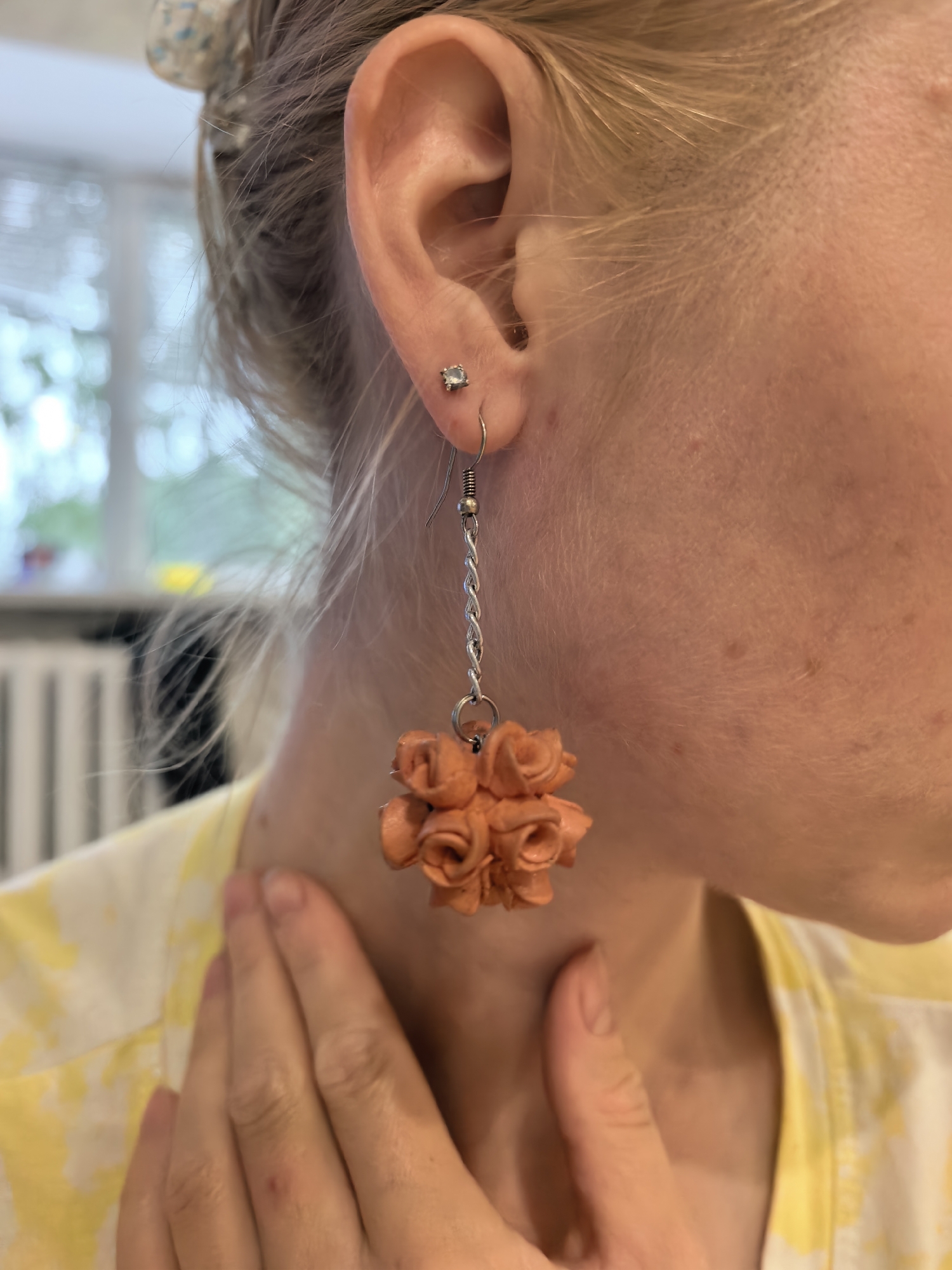 A woman's profile is shown, focusing on her right ear and neck. She is wearing an earring with a silver chain and a cluster of small, orange, rose-shaped charms. A silver stud earring is visible in her earlobe. Her hair is blonde and pulled back loosely. She is wearing a yellow and white tie-dye shirt. Her right hand is touching her neck. The background is blurred, showing what appears to be an indoor setting with a window and possibly blinds. There are no indications of Sterlitamak, Russia, in the image.