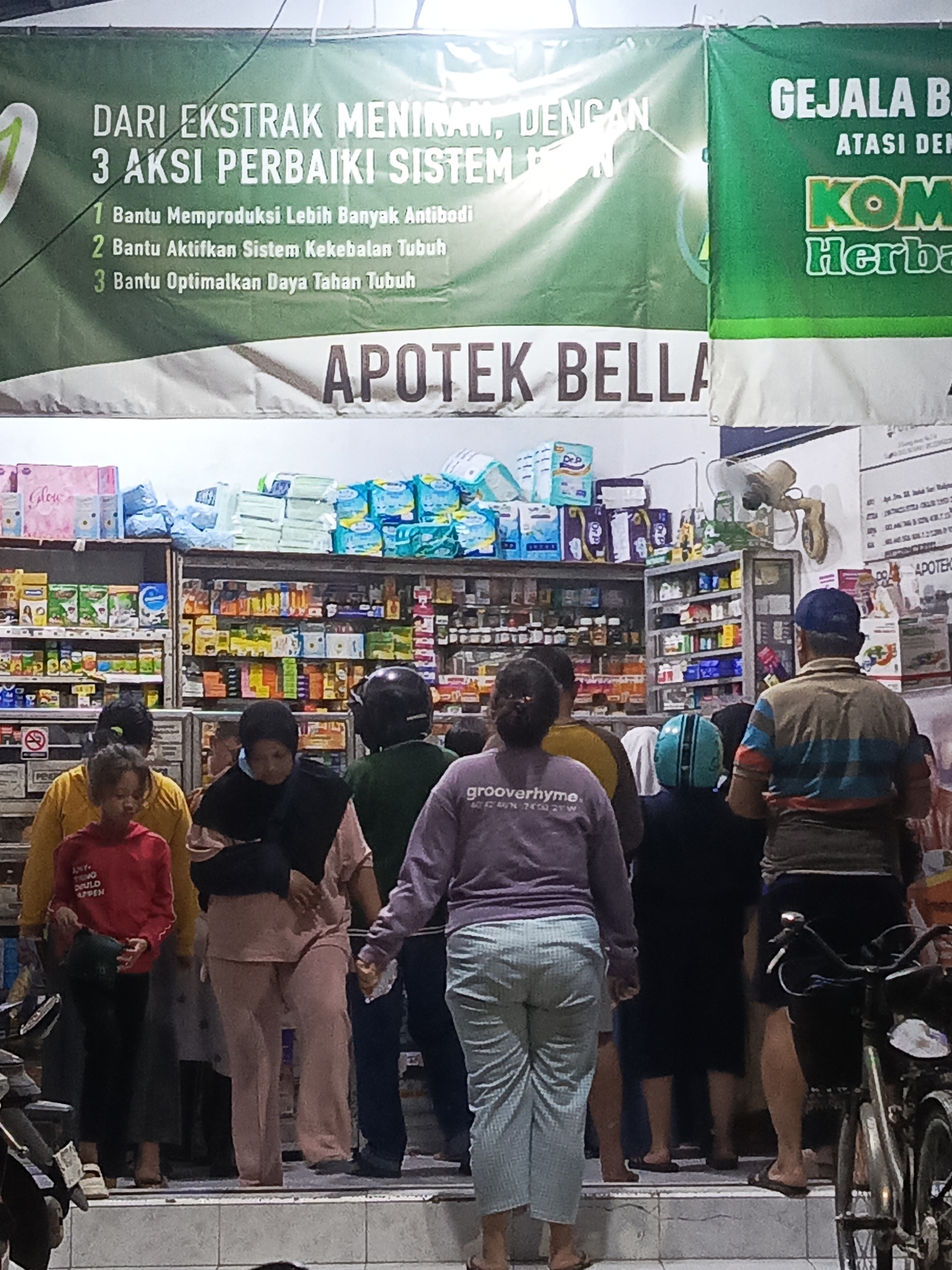 This is a photograph of a group of people standing outside an apotek (pharmacy) in Surabaya, Indonesia. The apotek appears to be named "APOTEK BELLA". A large green banner hangs above the storefront, with text in Indonesian. On the left side of the banner, it reads "DARI EKSTRAK MENIRAN, DENGAN 3 AKSI PERBAIKI SISTEM IMUN". Below this, three points are listed: "1. Bantu Memproduksi Lebih Banyak Antibodi", "2. Bantu Aktifkan Sistem Kekebalan Tubuh", and "3. Bantu Optimalkan Daya Tahan Tubuh". On the right side of the banner, it reads "GEJALA BEB" and "ATASI DEN", with the word "KOM" prominently displayed in large, green lettering, followed by "Herba".
In the foreground, several individuals are visible, some facing away from the camera. One person is wearing a purple hooded sweatshirt with "grooverhyme." printed on it. Another person is wearing a yellow jacket, and a child in a red hoodie is to their left. Further back, a person in a pink outfit is standing. Behind the people, shelves are stocked with various pharmaceutical products, including boxes of diapers and bottles. A bicycle is parked on the right side of the frame. The scene is lit by artificial lights, creating a somewhat dim environment.