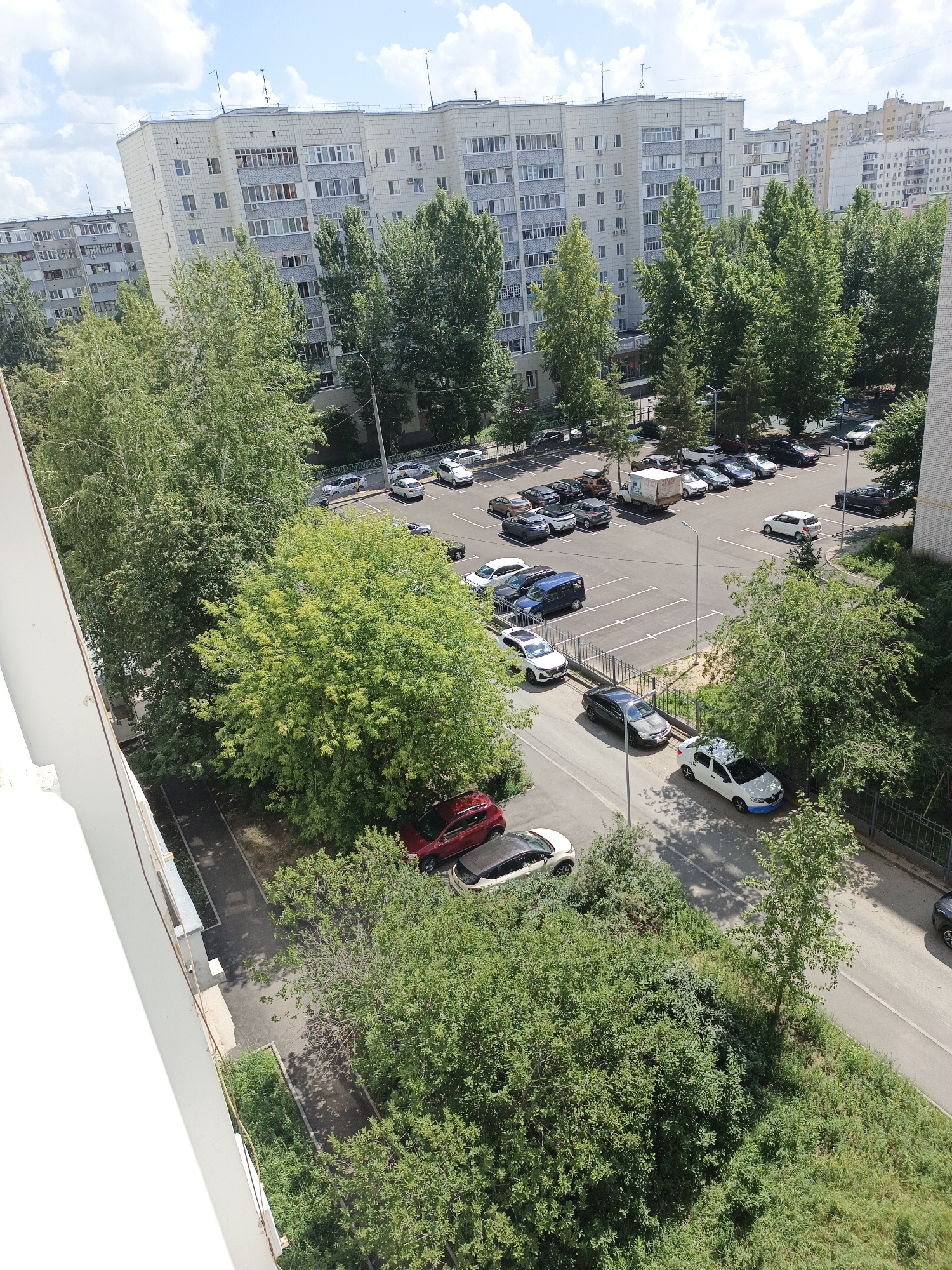 The image captures an aerial view of a residential area in Kazan, Russia, on a bright, partly cloudy day. In the background, several multi-story apartment buildings, characteristic of Soviet-era architecture, are visible. These buildings are light-colored with numerous windows and balconies. A prominent feature is a paved parking lot filled with numerous cars of various makes and models, parked in designated spaces. Green trees, with lush foliage, are interspersed throughout the scene, bordering the parking lot and the buildings. A road runs along the edge of the parking area, with a sidewalk and more greenery. The overall scene depicts a typical urban residential environment with ample parking and vegetation.