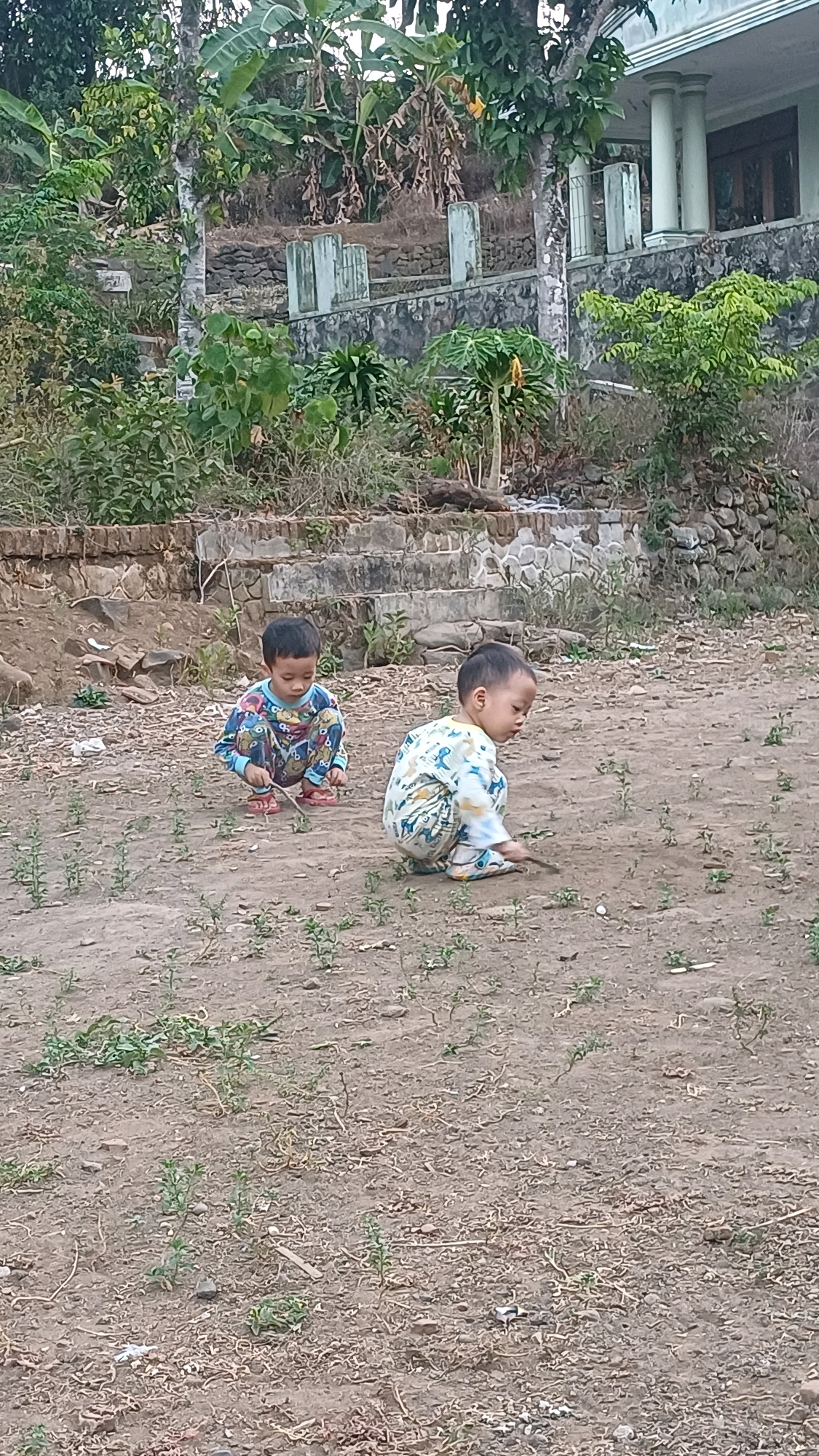Two young children are crouched on a dirt ground outdoors. The child on the left, wearing pajamas with a blue, pink, and yellow pattern, is holding a small stick and looking down. The child on the right, in light-colored pajamas with a green and yellow pattern, is also holding a stick and appears to be drawing in the dirt. The background shows a hillside with various plants, stone steps, and a building with white pillars. This scene is located in the village of Wonokerso, Indonesia.