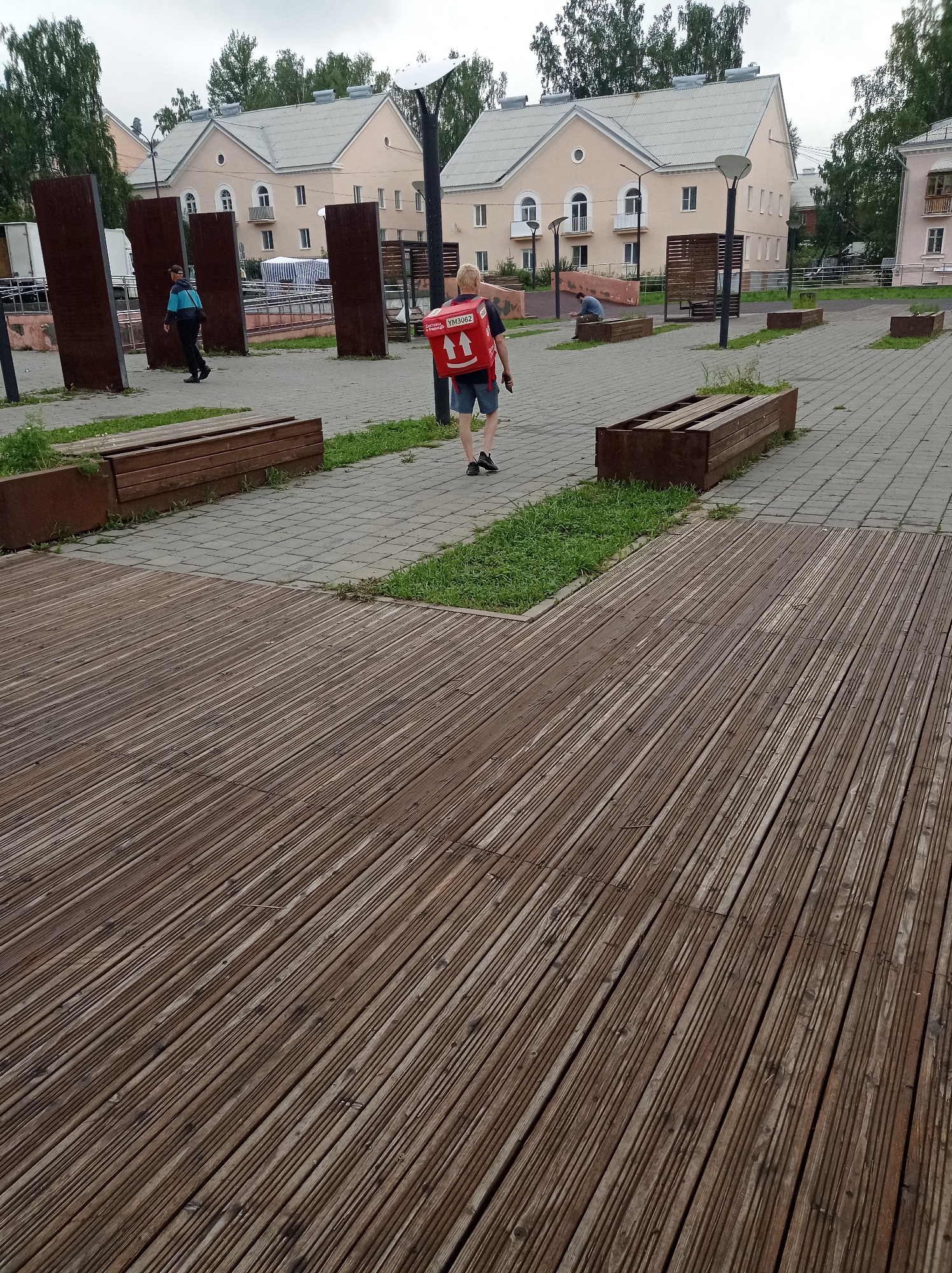 The image depicts an outdoor public space in Degtyarsk, Russia, likely a park or plaza. In the foreground, a weathered wooden deck occupies the right half of the frame, its planks running diagonally. To the left of the deck, a paved area with a geometric pattern is visible, featuring several long, rectangular planters made of dark metal and wood, some with grass growing in them.
In the middle ground, a person wearing a red delivery backpack with the logo "Яндекс Еда" (Yandex Food) is walking away from the camera towards the right. They are wearing dark shorts and a dark t-shirt. To the left, another person in a blue and black uniform with a cap is walking towards the left. In the background, two-story apartment buildings with beige facades and grey roofs are visible, interspersed with trees. Several dark, vertical metal structures are also present in the middle ground. The sky is overcast, suggesting a cloudy day.