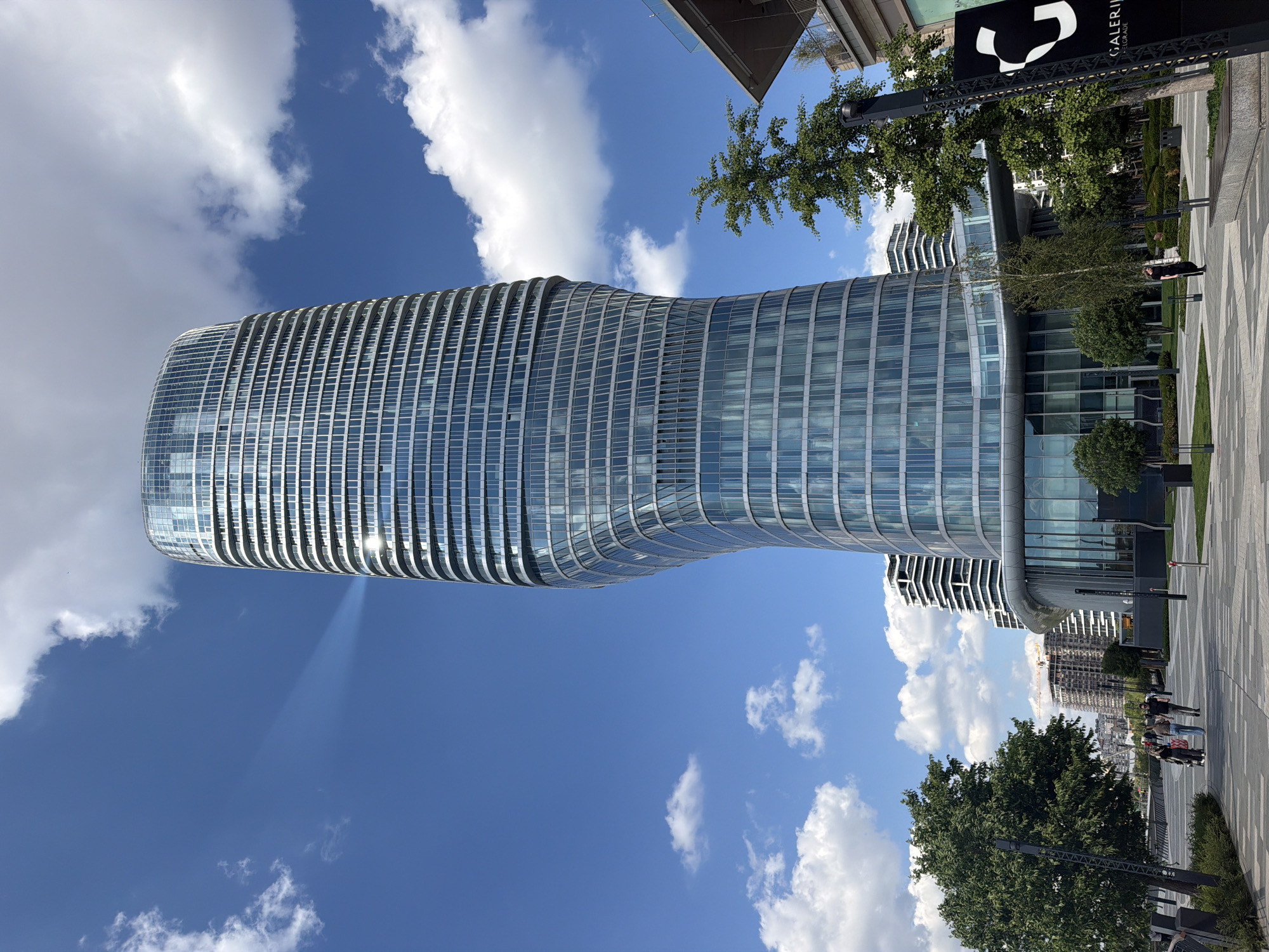 The image shows a modern, glass-clad building against a blue sky with scattered clouds. The building has a distinctive, curved shape, narrowing in the middle. It is located in Belgrade, Serbia, as indicated by the "Galerija" sign in the background. A sidewalk with a patterned surface is visible in the foreground, with sparse vegetation and trees bordering the building. People can be seen walking along the sidewalk in the distance. The image appears to have been rotated to a vertical orientation.