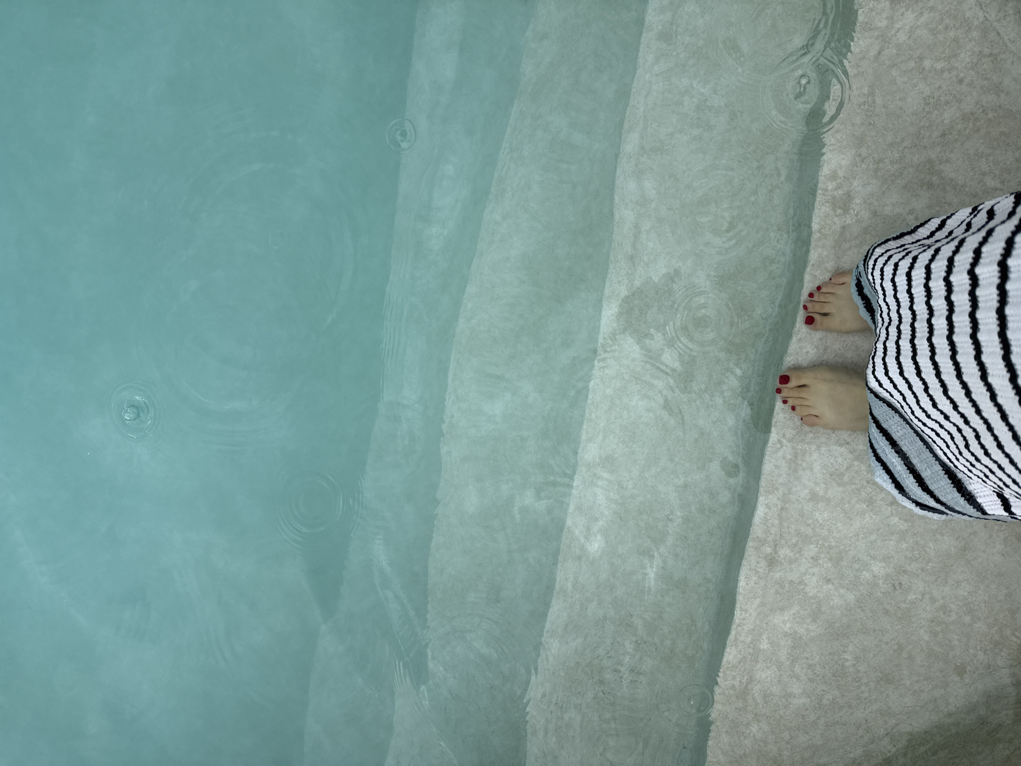 The image presents an aerial, downward-looking view of a person's bare feet at the edge of a tranquil body of water, likely a pool or shallow water feature. The person's feet, adorned with bright red painted toenails, are positioned on a light grey, textured surface to the right. A black and white striped garment, possibly a towel or robe, partially covers the lower legs.
The water, a serene light blue-green hue, dominates the left and central parts of the frame. Several broad steps, also made of the same light grey, textured material as the surrounding surface, descend gently into the water. The water's surface is not perfectly still; numerous small, concentric ripples are visible across both the open water and over the submerged steps, suggesting the gentle impact of raindrops or a slight disturbance. The lighting is soft and diffused, creating a calm atmosphere. No text is visible in the image.