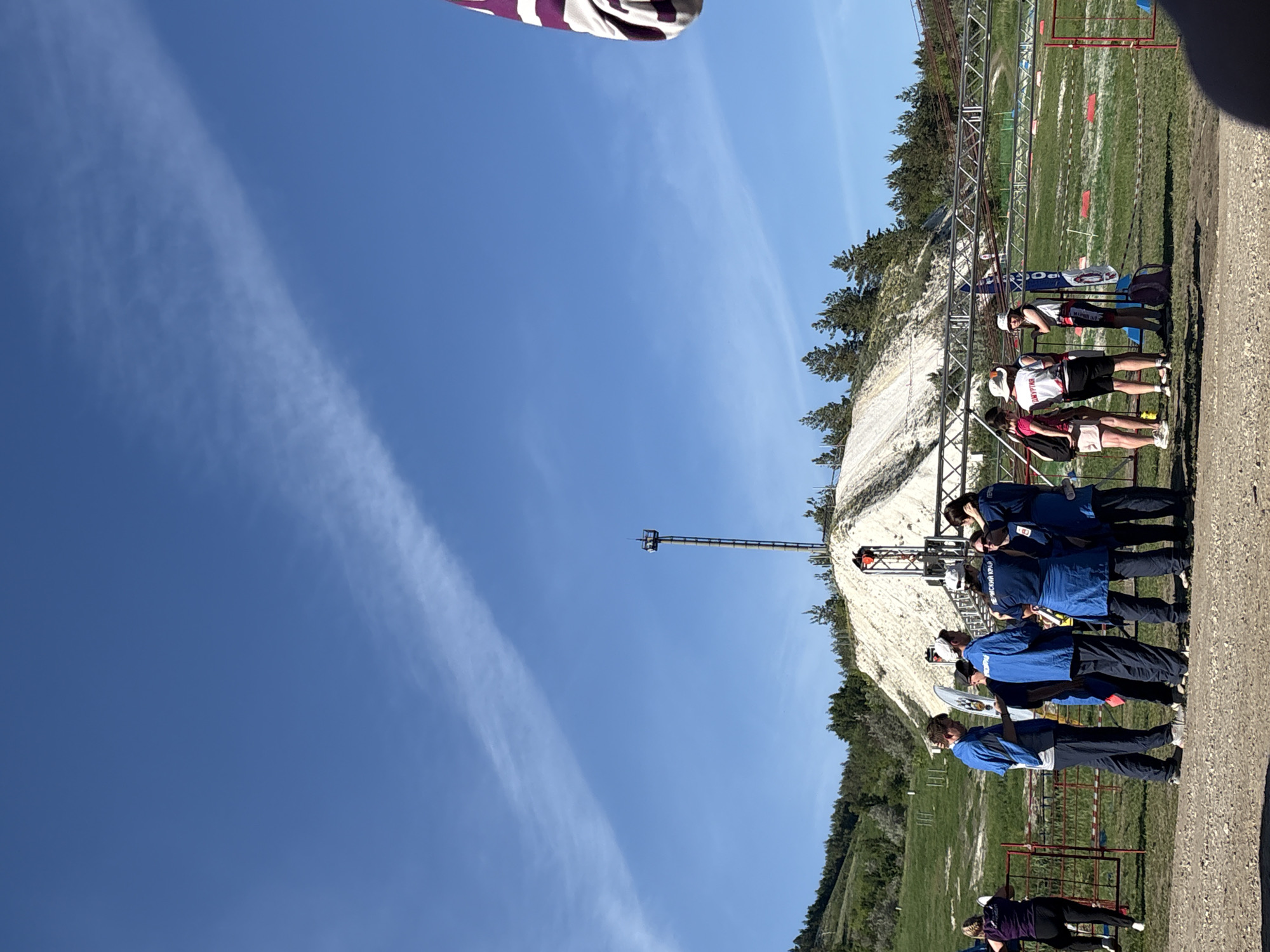 The image shows an outdoor scene in Khvalynsk, Russia. There are people standing near a ski jump structure built on a hillside. Some individuals are wearing blue tracksuits, possibly representing a team or organization. Other individuals are wearing casual athletic wear. The ski jump structure is made of metal scaffolding and leads to a grassy field marked with lines and targets. In the background, a steep, white, chalky hillside rises with sparse trees along the top. The sky is clear blue with a wispy cloud formation extending across the frame.