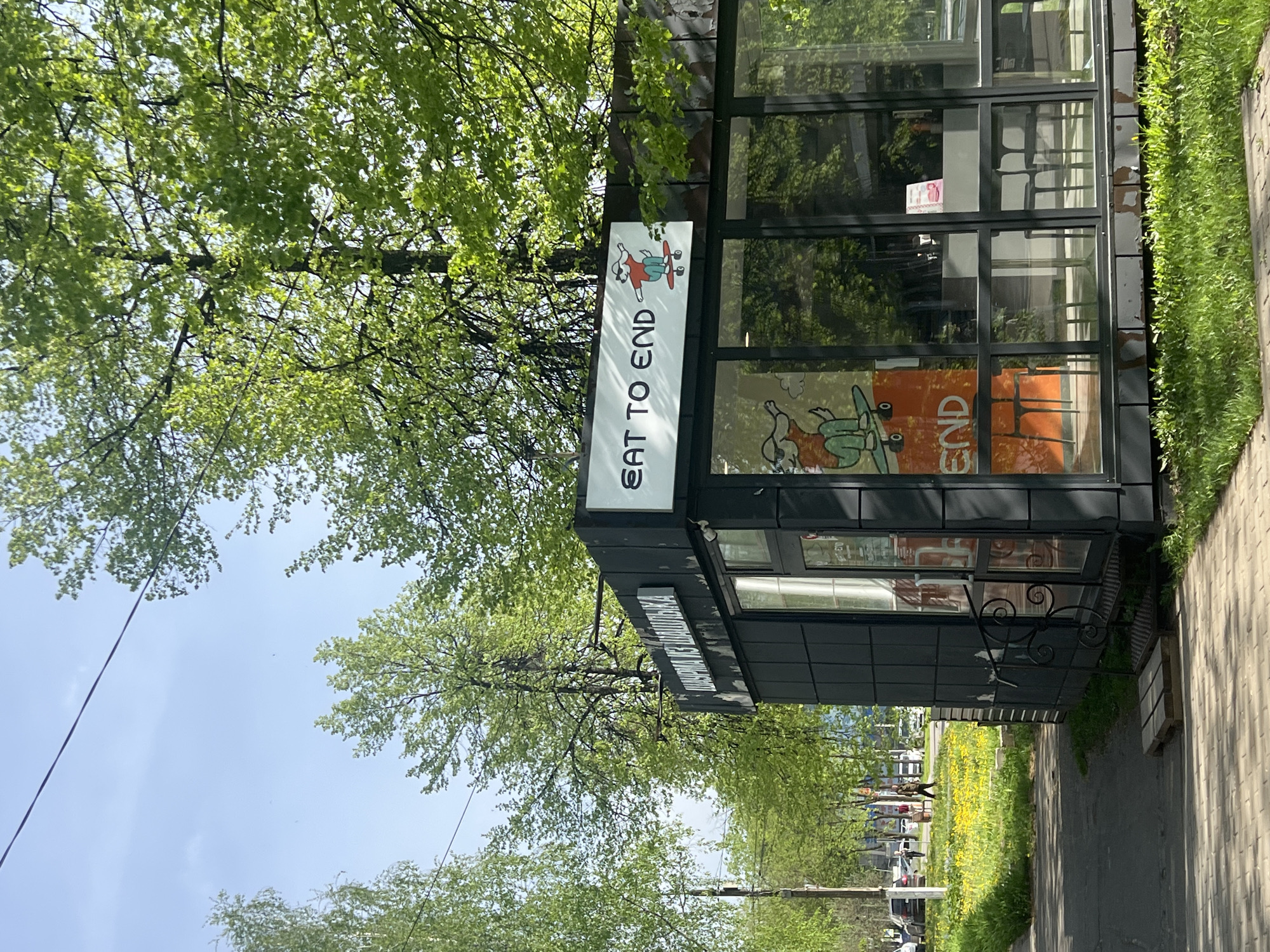 The image shows a food stand in Chaikovsky, Russia. The stand has a sign that reads "EAT TO END" with a cartoon skateboarder. The stand is dark gray with black framed windows. The windows have a cartoon image. The stand is located on a street with trees and a sidewalk.