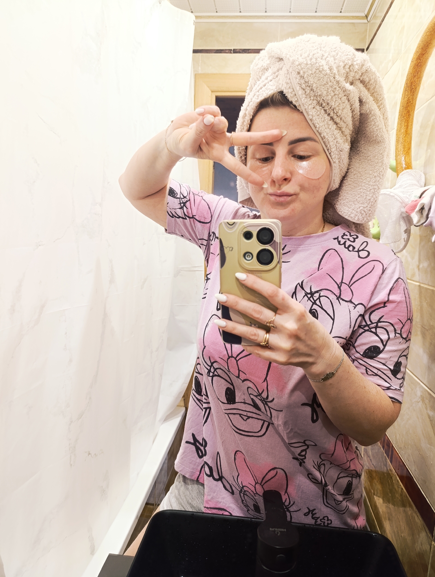 A woman is taking a mirror selfie in what appears to be a bathroom, engaging in a personal care routine. She has a light beige terry cloth towel wrapped around her head and wears translucent pinkish-orange eye patches under her eyes. Her attire consists of a casual light pink t-shirt adorned with black outline illustrations of Disney's Daisy Duck characters and bow motifs, with grey shorts partially visible.
She holds a light gold-colored smartphone, featuring multiple prominent camera lenses, in her left hand to capture the image. Her fingers are decorated with gold-toned rings, and a delicate gold chain bracelet is on her right wrist. With her right hand, she forms a "peace sign" gesture near her left eye, while her facial expression is a playful pout.
The background on the left features a plain white wall or shower curtain, contrasting with the beige-brown ceramic tiles on the right. A dark-colored, modern bathroom sink and faucet are visible in the foreground; the faucet bears faint white text, "HERTUSK". A partial wooden door frame and various hanging bathroom items, such as towels or sponges, are also discernible. The scene conveys a relaxed, intimate moment of self-care.
Visible text includes "HERTUSK" on the faucet and stylized, partially obscured text on her t-shirt, which clearly features the recognizable Disney character Daisy Duck.