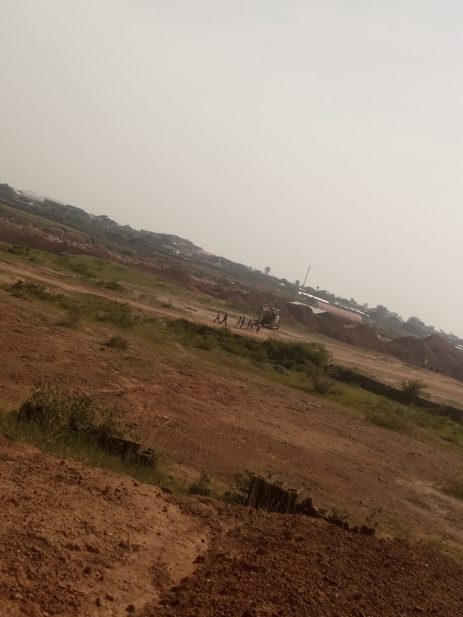 The image depicts a dusty, open terrain with scattered vegetation and some construction activity. The ground is predominantly red-brown soil, indicative of arid or semi-arid conditions. In the mid-ground, there is an excavator, a heavy machinery used in construction, accompanied by a group of people. Further in the background, there are several structures, likely houses or buildings, suggesting a developing settlement or a rural area. The overall environment appears to be an outdoor scene under a hazy sky. The tilt of the camera suggests the image was taken on a slope or at an angle. This scene is located in Kwang, Nigeria.