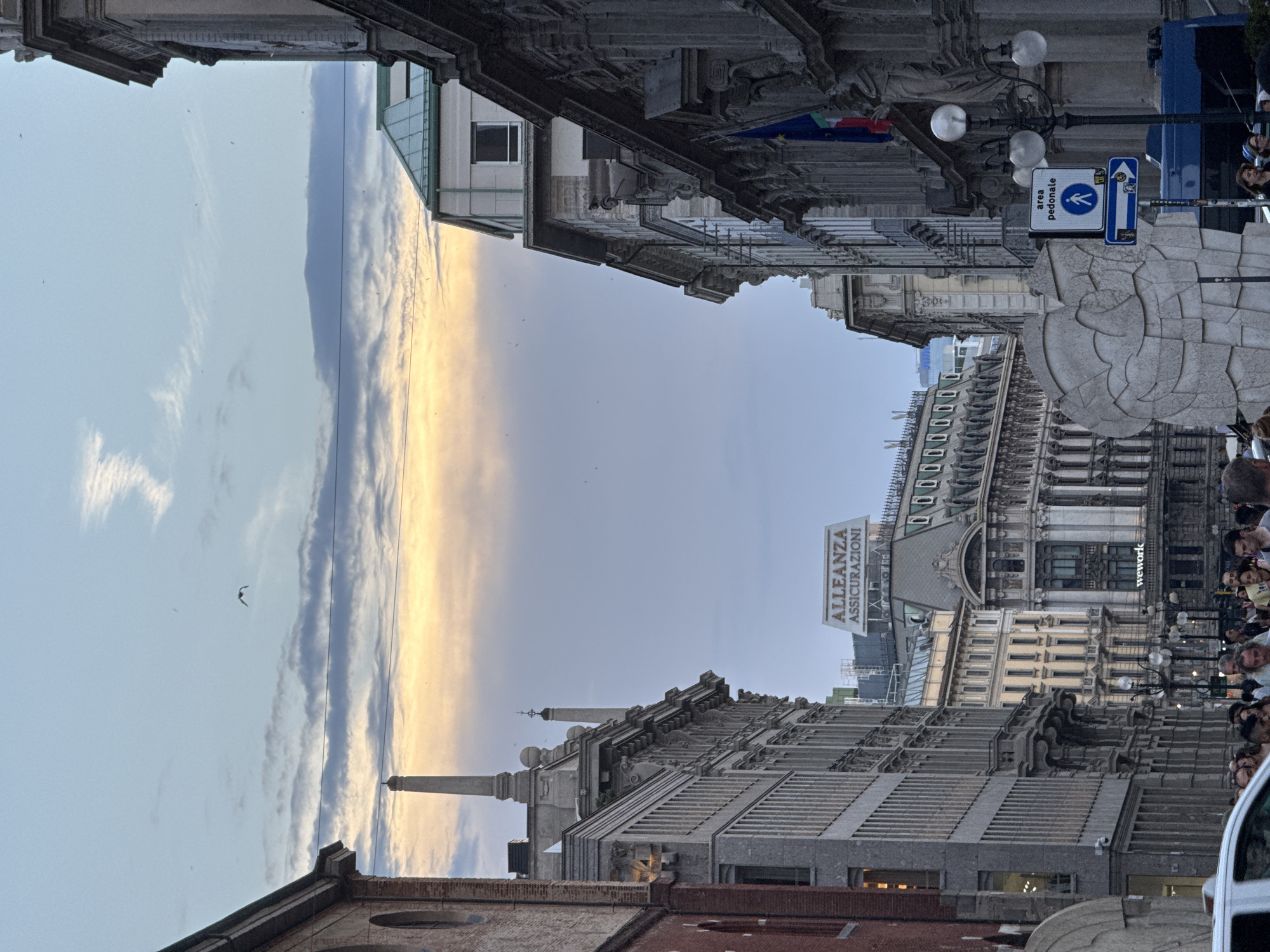The image depicts a view of a European city street, likely Milan, Italy, during dusk. The sky is a blend of light blue and orange hues, with scattered clouds and several birds in flight. Tall, ornate buildings line both sides of the street, showcasing a mix of architectural styles. On the left, a brick building is partially visible, followed by a grey stone building with multiple windows. On the right, a grander, darker stone building features intricate carvings and sculptures.
In the background, several other buildings are visible, including one with a sign that reads "ALLEANZA ASSICURAZIONI." Another building in the distance has a "wework" logo. A street lamp with multiple globes stands on the right side of the street, near a pedestrian zone sign. A large, abstract stone sculpture is prominent in the foreground on the left. A white car is partially visible at the bottom left of the frame. A crowd of people are gathered on the street, suggesting a public space or thoroughfare. The overall impression is one of urban life at the end of the day.
