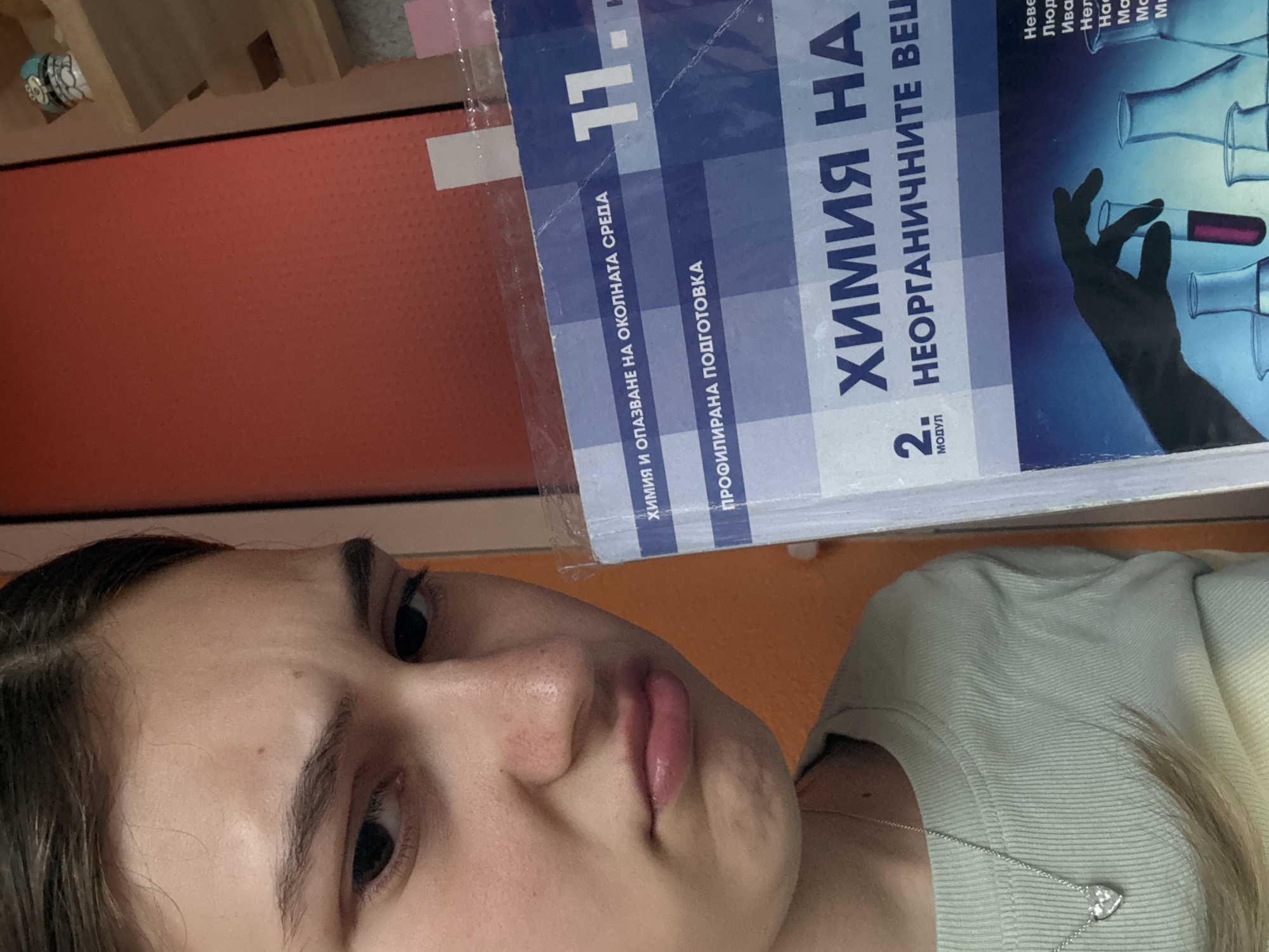 The image shows a young woman holding a chemistry textbook titled "ХИМИЯ НА НЕОРГАНИЧНИТЕ ВЕЩЕСТВА" (Chemistry of Inorganic Substances) for 11th grade. The textbook's cover features a hand in a lab glove holding a test tube with a colored liquid. The environment appears to be indoors, with an orange wall partially visible in the background. There are no indications that the image was taken in Panagyurishte, Bulgaria, or any information about the location.