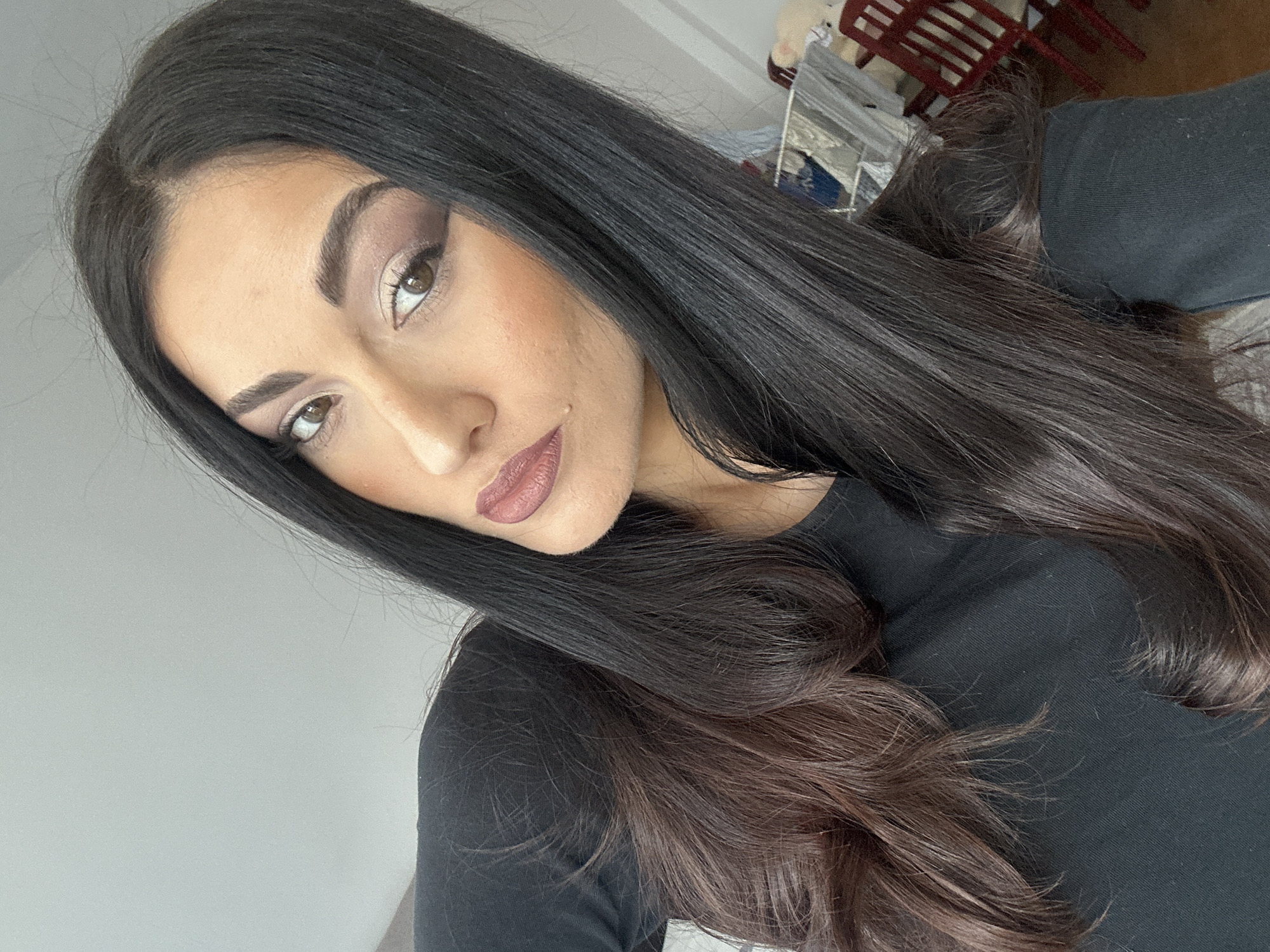 A medium-shot, slightly angled portrait of a woman. She has dark hair with brown highlights at the ends, fair skin, and brown eyes. Her makeup includes eyeshadow, blush, and lipstick. She is wearing a black, long-sleeved shirt. The background is a plain white wall, and there are some indistinct objects in the upper right corner of the frame, including what appears to be a red chair. The location and city are unknown.