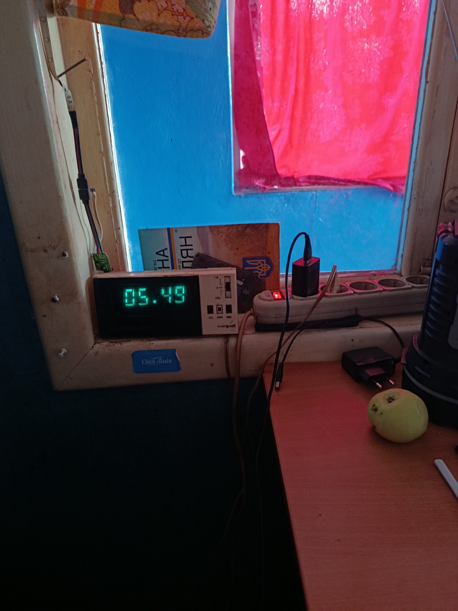 The image shows a close-up view of a window sill with various objects on it. A digital clock displays the time as 05:49. To the right of the clock is a power strip with a red indicator light on and two black power adapters plugged in. A small green apple is positioned on the right side of the frame. In the background, a bright pink curtain is visible through the window, along with a blue painted wall. A book with Cyrillic text is partially visible behind the clock. The overall scene appears to be an indoor setting, possibly a residential room, with a focus on electrical devices and everyday items. The image does not contain any identifiable landmarks or contextual clues that would place it specifically in Kremenchuk, Ukraine.