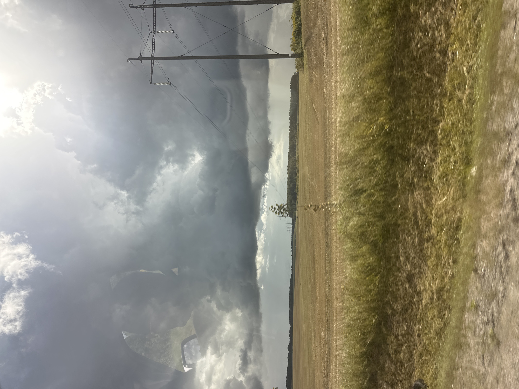 The image depicts a landscape scene with a field and a stormy sky. In the foreground, there is a blur of green and yellow vegetation, suggesting movement. The middle ground features a vast, flat field, appearing to be recently harvested or fallow, with a light brown color. In the distance, a line of trees forms a dark horizon.
Dominating the upper portion of the image is a dramatic sky filled with dark, ominous storm clouds. Sunlight pierces through the clouds in some areas, creating a contrast between light and shadow. Two tall, gray electricity pylons with wires stretching between them are prominent on the right side of the frame. There is a visible reflection on the left side of the image, likely from a window, showing indistinct shapes and the sky. The overall impression is one of approaching or passing weather. The image is taken from a moving perspective, possibly from a vehicle.