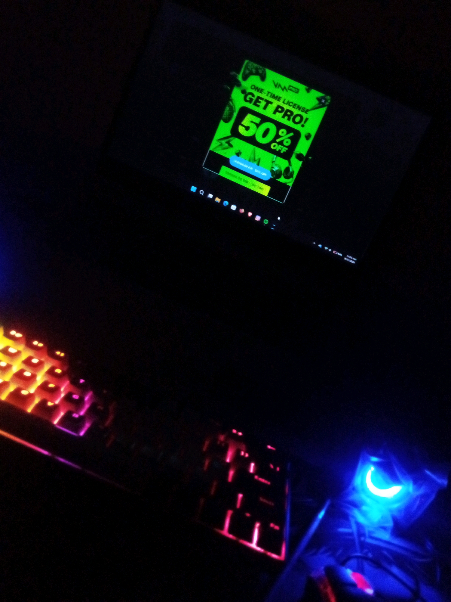 In a dimly lit room, likely a home office or gaming setup, a computer monitor displays a bright green advertisement for "VNN PRO" offering a "ONE-TIME LICENSE" for "50% OFF". The advertisement features various gaming icons. Below the monitor, a keyboard with RGB backlighting is visible, emitting red, orange, and purple hues. To the right of the keyboard, a gaming mouse with blue and pink glowing accents is partially visible. The overall ambiance suggests a nighttime setting, possibly in Boyoteluk, Indonesia, given the context of the prompt.