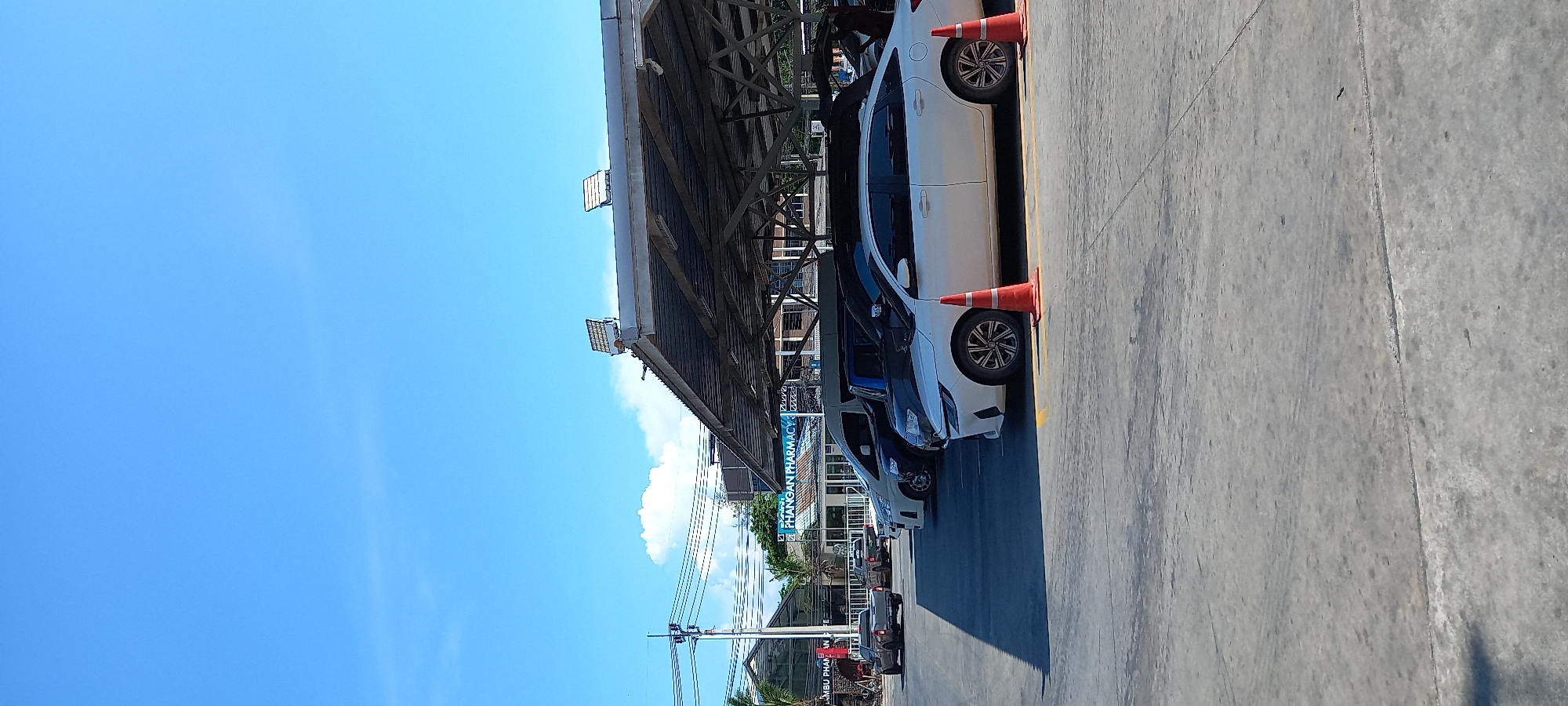 This image captures a bright, sunny daytime scene in what appears to be a commercial area or parking lot in Moo 1, Thailand. The sky is a clear, vibrant blue with a few scattered white clouds.
In the foreground, a concrete or asphalt surface serves as a parking area, marked with yellow lines. Several cars are parked parallel to the viewer, predominantly sedans; a white car with distinctive black and silver alloy wheels is most prominent, accompanied by at least one darker-colored vehicle. Orange traffic cones are placed next to the wheels of the white car.
An overhead structure, resembling a large metal-framed canopy or the underside of a building's extension, provides shade over the parked vehicles. Utility poles and power lines crisscross the mid-ground, indicating a developed streetscape.
In the background, beyond the parked cars and utility infrastructure, other buildings and tropical greenery are visible. A clear blue sign with white lettering is prominent, reading "PHANGAN PHARMACY," strongly suggesting the location is Koh Phangan, Thailand. Below this, another partially visible sign appears to begin with "KO SAMUI PHAR," possibly indicating another branch or related business.
The scene depicts a calm, ordinary moment with no people visible or specific events taking place. The weather is clear and bright, characteristic of a sunny day.