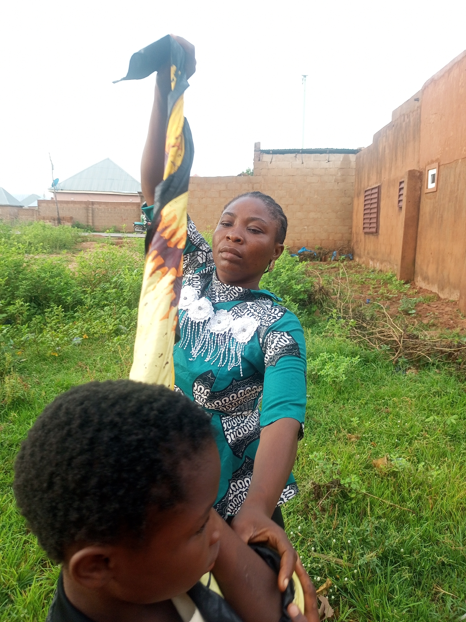 In Tempure, Nigeria, a woman wearing a teal and white patterned shirt holds up a yellow and black patterned cloth. She is outdoors, with a child's head in the foreground, partially obscuring the woman's body. Behind them, a brick wall and a house with a tan exterior and a barred window are visible. The ground is covered in green grass and sparse vegetation. The sky is overcast.