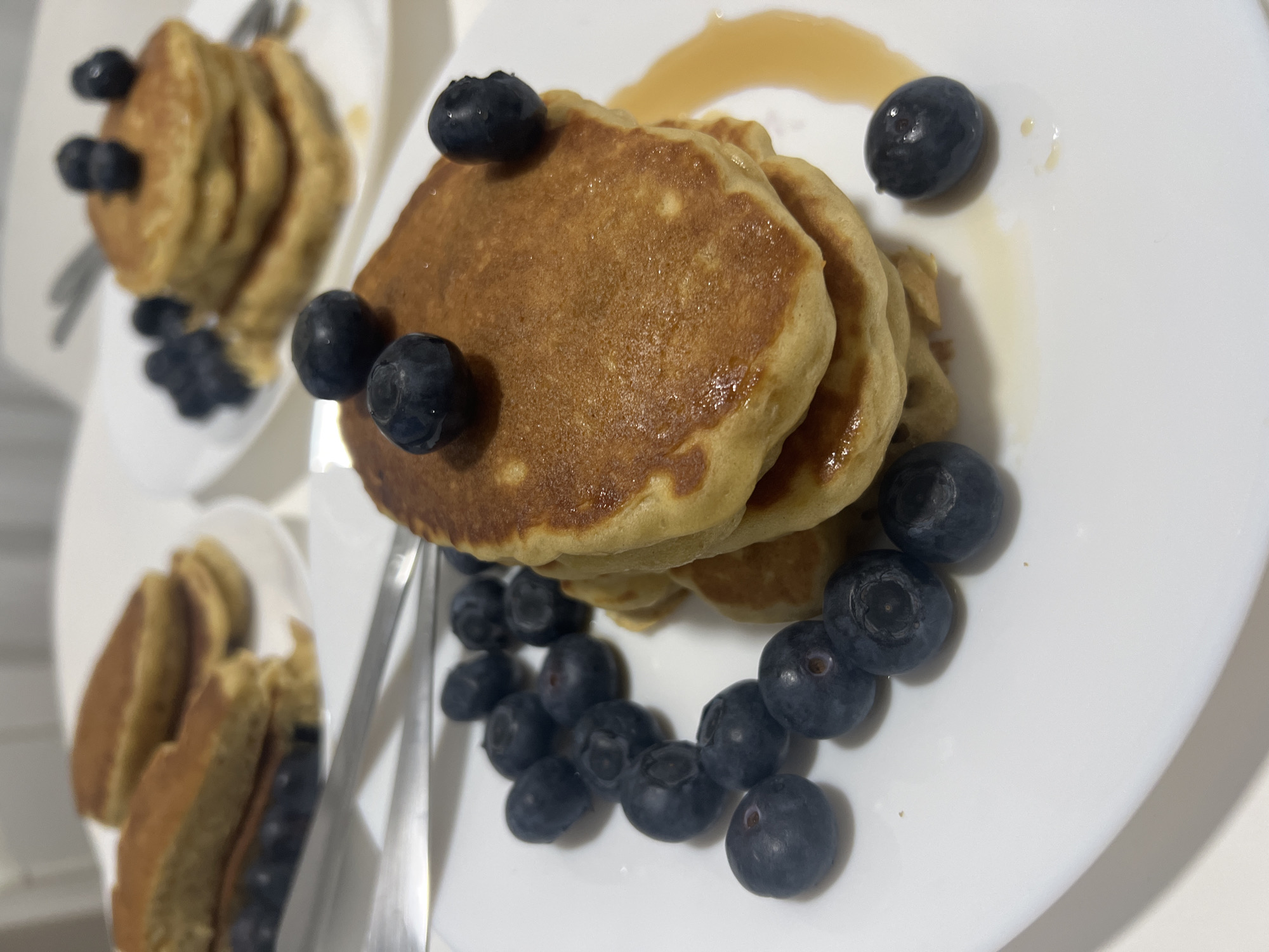 The image shows three white plates of pancakes garnished with blueberries. Each plate has a stack of pancakes and a scattering of blueberries. Syrup is drizzled on one plate. Silverware is visible near the pancakes. The setting is a bright, indoor location. There is no indication of Mogán, Spain in this image.