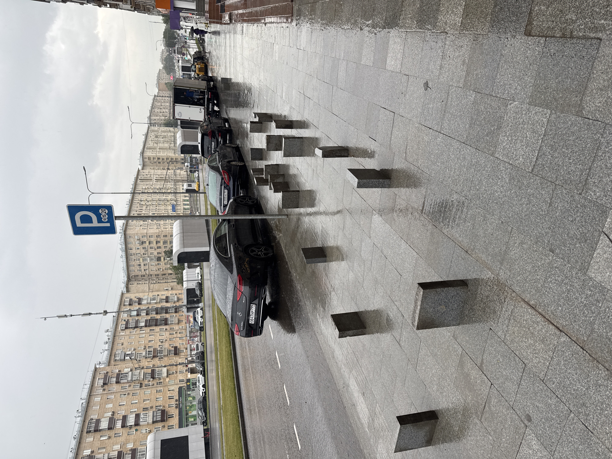 On a rainy day in an unknown city, a street scene unfolds with apartment buildings lining one side and a sidewalk with parked cars on the other. The street is wet, reflecting the overcast sky. Several cars, including a black Mercedes-Benz, are parked along the curb. Further down the street, a delivery truck is being unloaded by at least one person. A few other vehicles are visible, including a yellow loader. A person with an umbrella walks on the wet sidewalk. The sidewalk is paved with grey tiles and features several short, rectangular granite bollards. In the foreground, a pattern of these bollards is arranged in a semi-circular shape.