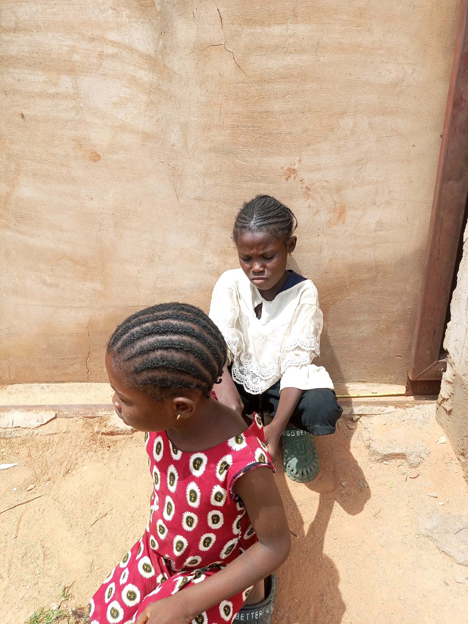 Two young girls are sitting on the sandy ground in front of a rough, textured, tan-colored wall. The wall appears to be made of plaster or stucco and has some visible cracks. To the right of the frame, a vertical metal pipe or post is attached to the wall. The girl in the foreground is wearing a red dress with a white and black circular pattern. She is looking to her left, and her hair is braided in neat rows. The second girl is seated behind the first, looking down with her eyes closed. She is wearing a white, lacy-sleeved blouse and black pants. She is wearing green clog-style shoes. The scene is illuminated by bright sunlight, casting shadows on the ground. The image was taken outdoors in a location that appears to be in a neighborhood in Tempure, Nigeria.