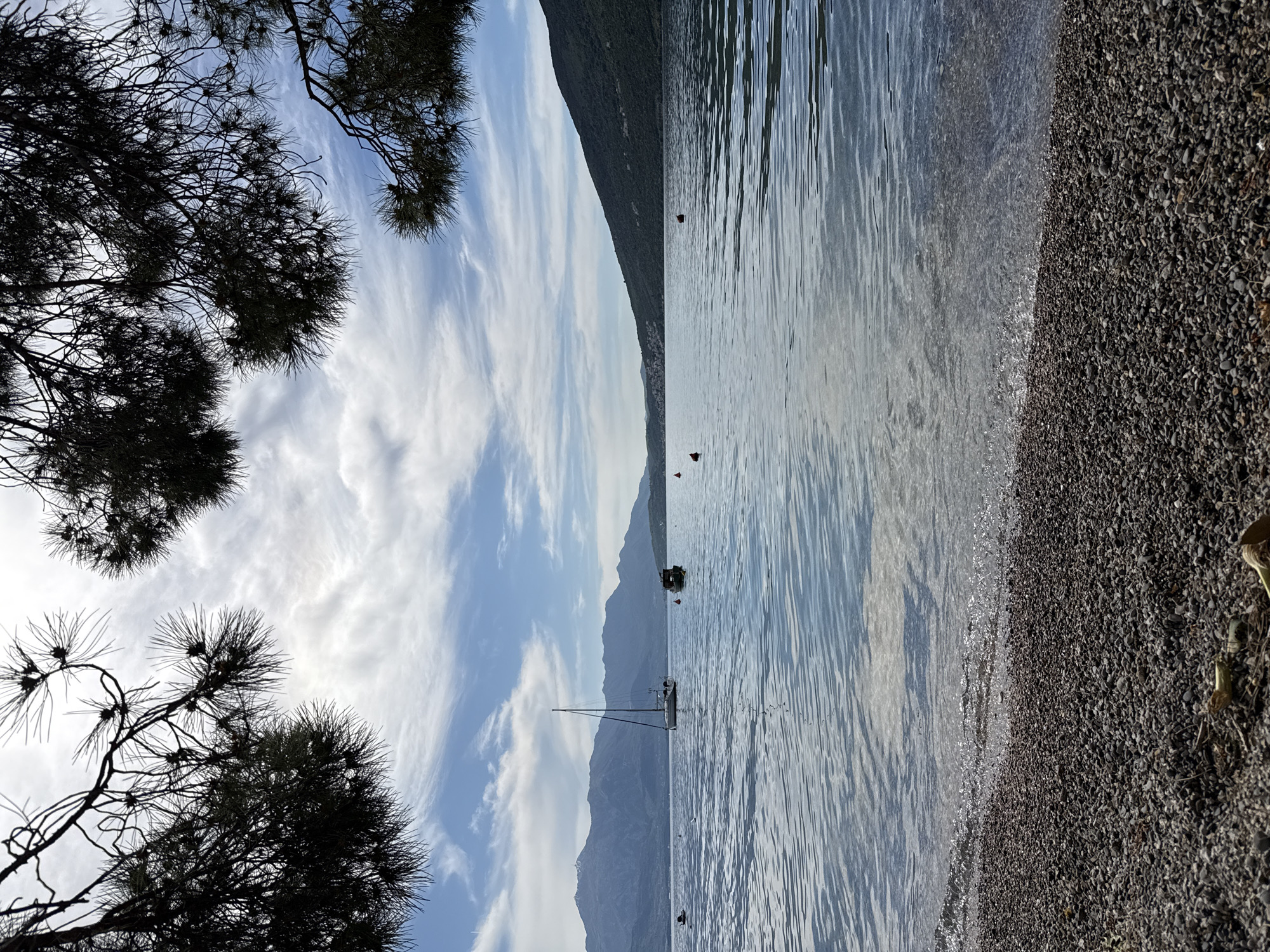 The image captures a coastal scene in Đenovići, Montenegro. The foreground features a pebbled beach meeting calm, rippling water. In the water, two boats, a sailboat and a smaller motorboat, are anchored. The background showcases a hazy mountain range stretching across the horizon beneath a sky filled with wispy clouds. Pine tree branches frame the upper left corner of the image. The lighting suggests it is daytime, with a gentle, diffused sun.