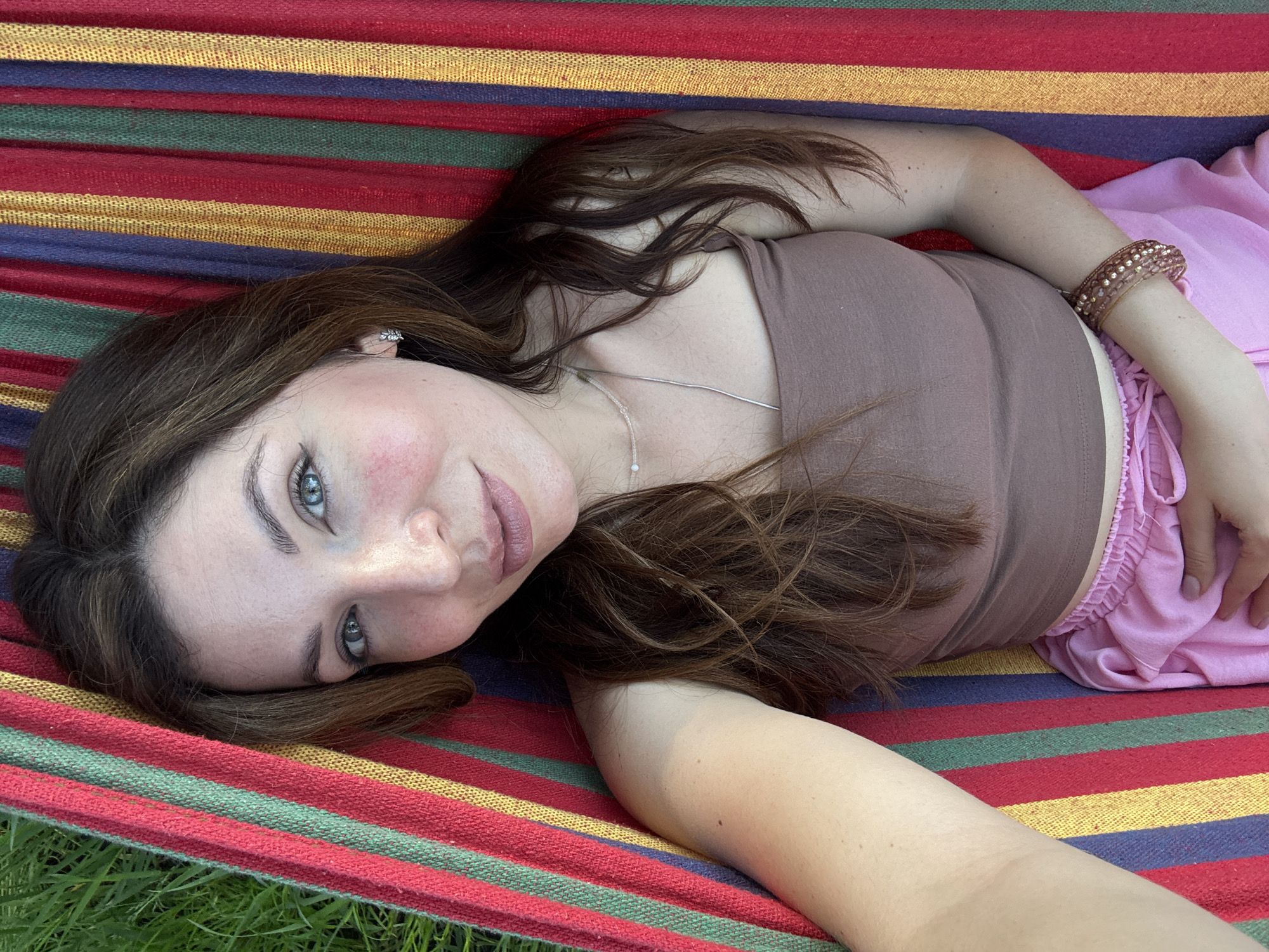 A woman is lying in a striped hammock outdoors. She is wearing a brown top and pink shorts. She has brown hair and blue eyes, and is wearing a necklace and a bracelet. The hammock is suspended over grass. The location is unknown.