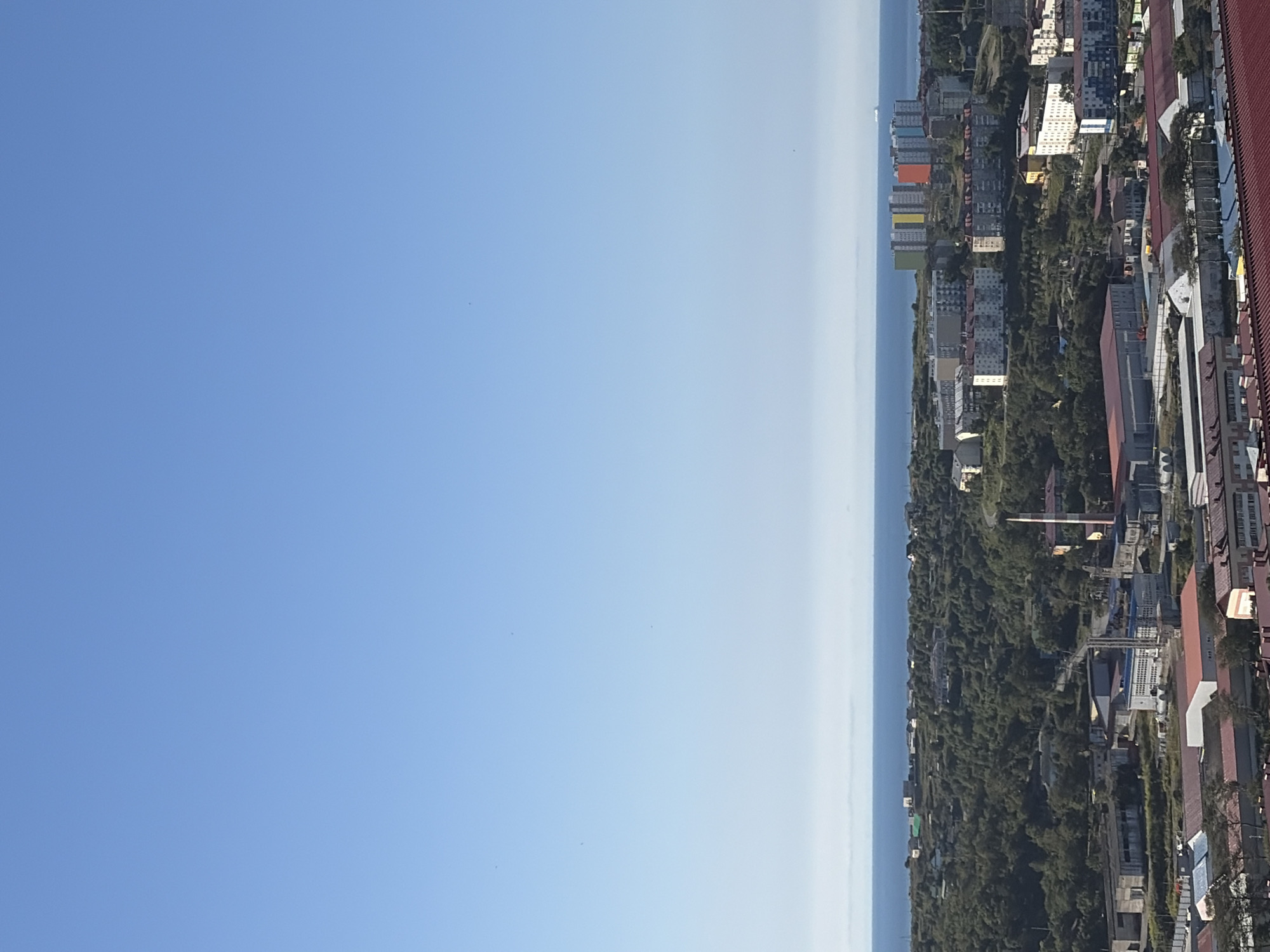 The image depicts a panoramic view of a coastal city, likely Korsakov, Russia, on a clear, sunny day. The scene is divided into distinct horizontal bands: a vast, pale blue sky dominates the upper half, transitioning to a hazy, light-colored horizon line that separates the sky from the sea. The sea itself is a deep blue, stretching across the frame, with a faint white line suggesting distant land or a fog bank.
Below the horizon lies a densely forested hillside, dotted with various buildings of different sizes and architectural styles. The trees are predominantly green, indicating a temperate climate. As the view descends, more urban structures become visible. These include residential buildings, some appearing to be multi-story apartment complexes, with varied color palettes. Notably, a cluster of brightly colored apartment buildings, featuring facades in yellow, green, orange, and blue, stands out against the otherwise muted tones of the cityscape. Industrial or commercial buildings with large, flat roofs are also present in the foreground, some with red roofing material. A tall, red and white striped industrial chimney is also visible among the buildings. The overall impression is of a coastal settlement with a mix of residential, industrial, and natural elements. No people are visible in the image.