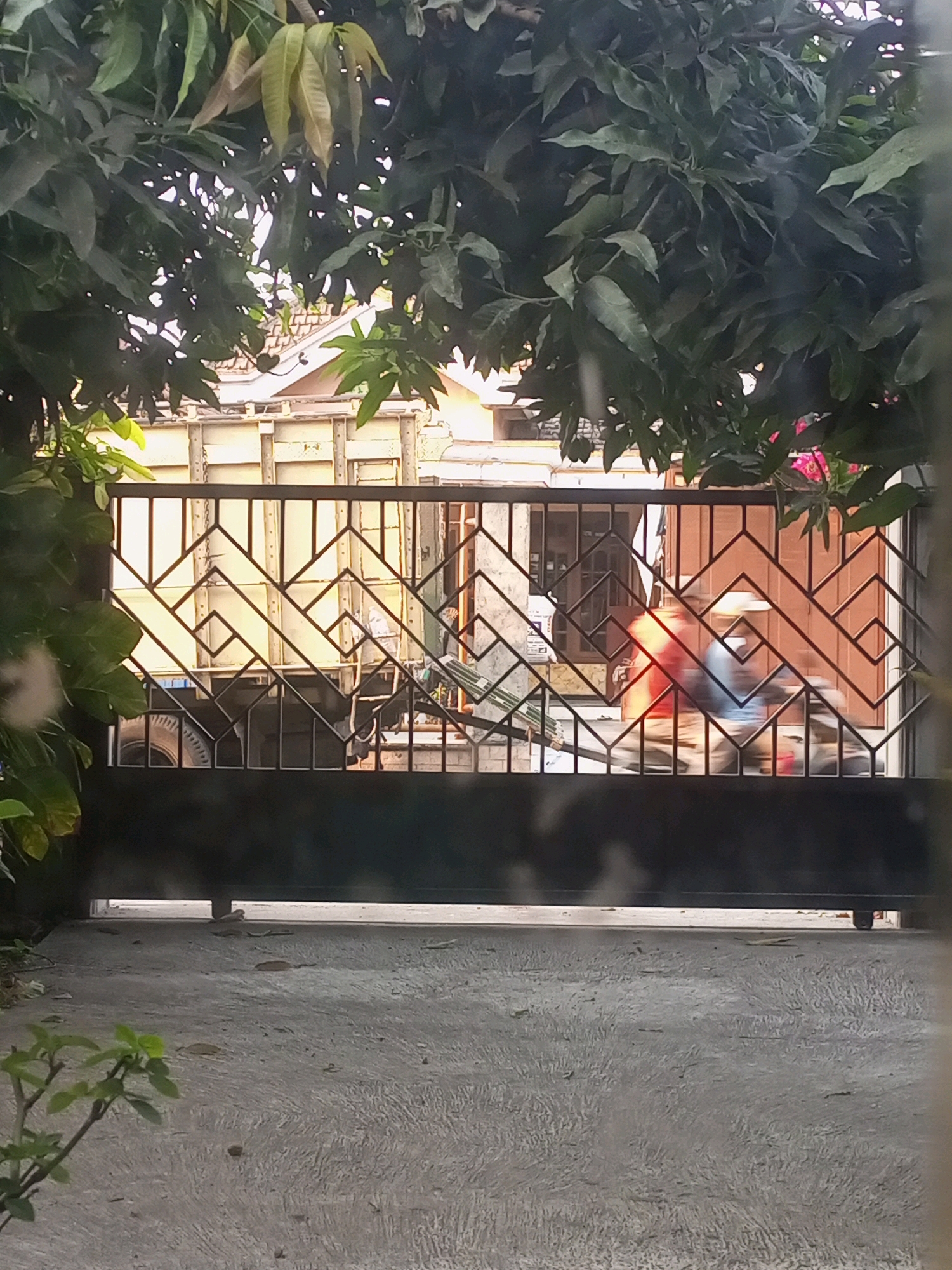 A gate with a geometric design made of dark metal bars stands in the foreground, partially obscuring a street scene. Behind the gate, a light-colored truck with wooden sides is visible. Two people on a motorcycle are blurred in motion, passing through the gate. The environment appears to be an outdoor setting, possibly a residential street in Ngasem, Indonesia, with buildings and foliage visible in the background. The ground in front of the gate is paved with concrete.