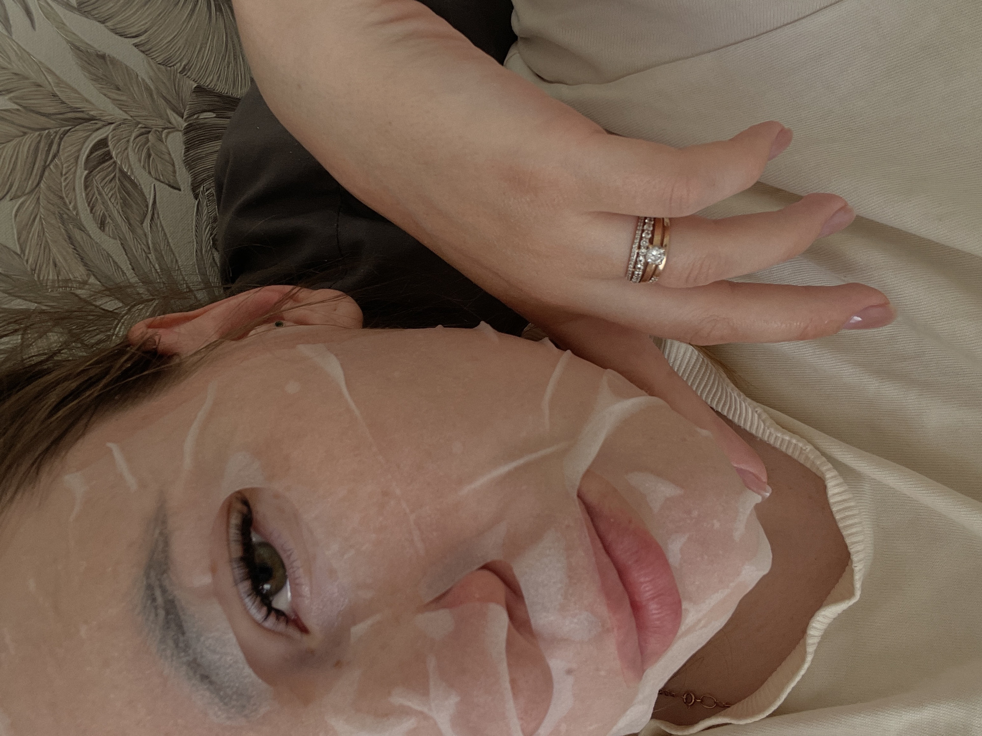 A fair-skinned person is lying on their back, with a sheet mask covering their face. Only the left eye, eyebrow, and lips are visible beneath the mask. The person's right hand is raised, displaying several rings on their ring finger, including what appears to be an engagement ring. They are wearing a cream-colored sweater. The setting includes a backdrop of a leafy pattern and what seems to be part of a dark-colored top. There is no evidence to indicate that the picture was taken in Chaikovsky, Russia.