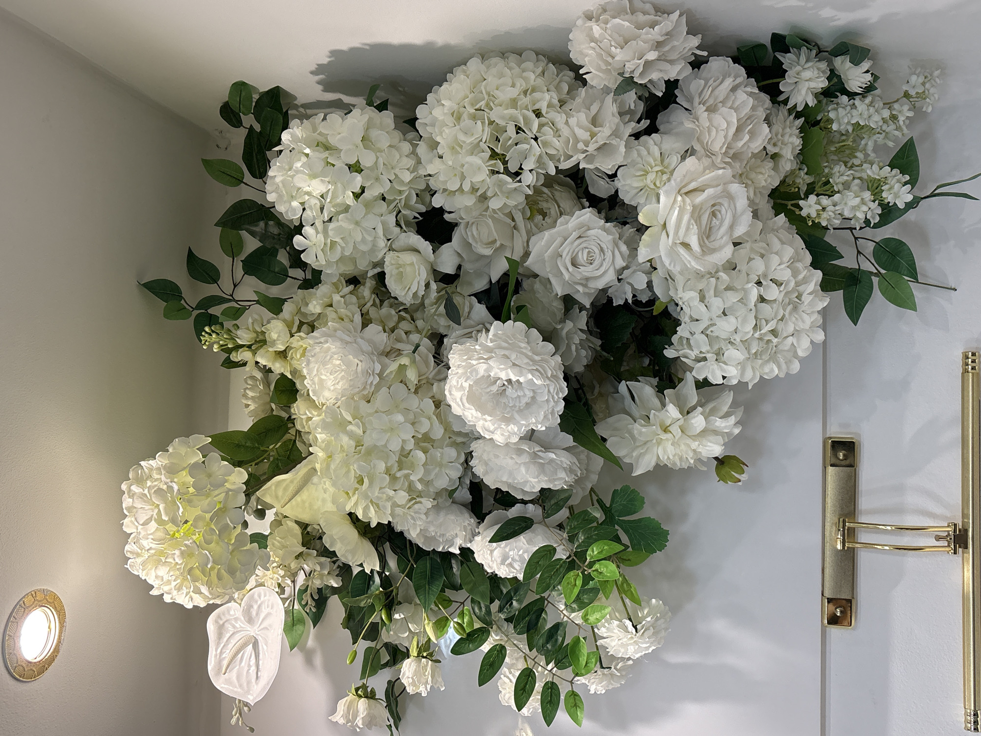 A large, opulent floral arrangement composed entirely of white flowers and greenery is displayed on a white wall. The arrangement features various types of flowers, including hydrangeas, roses, and anthuriums, with dark green leaves interspersed throughout. At the top left of the arrangement, a ceiling light fixture with a decorative, metallic trim is visible, casting a warm glow. Below the floral display, a golden picture light fixture is mounted horizontally on the wall. The wall itself appears to be a smooth, matte surface. The scene is well-lit, highlighting the textures and delicate details of the flowers. The location is a white-walled interior space, possibly a shop, gallery, or private residence. There are no people or animals present in the image. The composition is a still life of the floral arrangement and lighting fixtures.