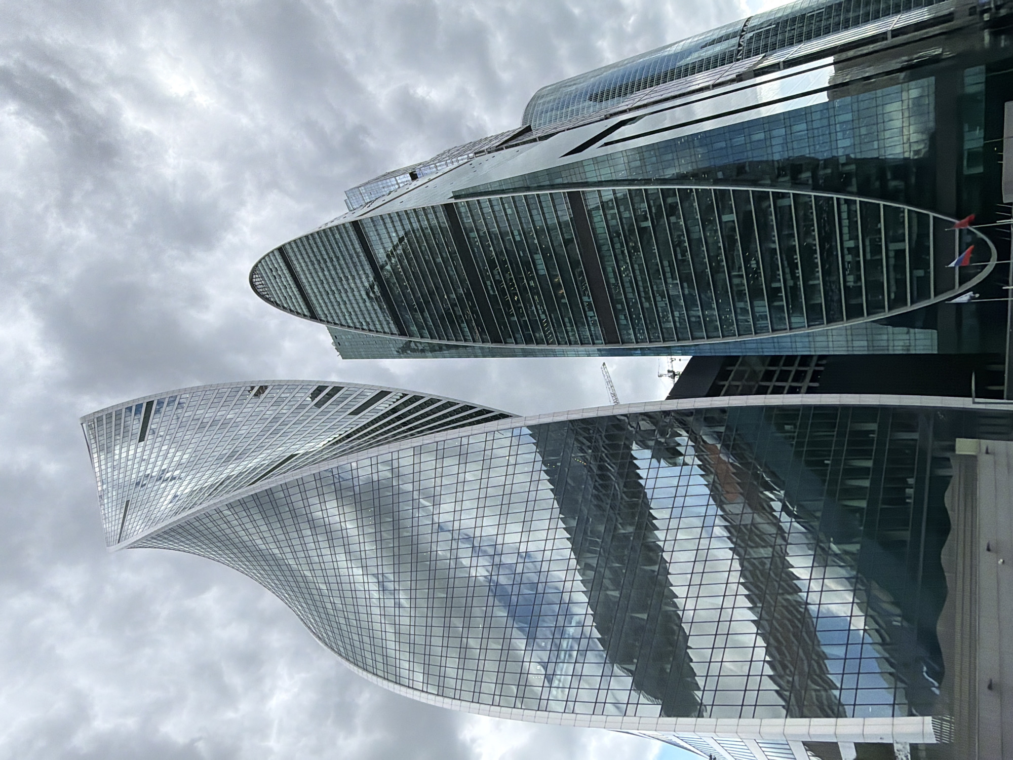 The image captures the architectural marvel of Moscow's modern skyline. Two prominent skyscrapers dominate the view against a backdrop of overcast skies filled with gray clouds. The lower building features a twisting design with a glass facade reflecting the cloudy sky. Above it, another skyscraper exhibits a streamlined, oval shape, partially obscured by the foreground structure. The Russian flags are visible near the second building. The photograph focuses on the buildings' design, emphasizing their modern architecture, and their integration into the urban landscape of Moscow.
