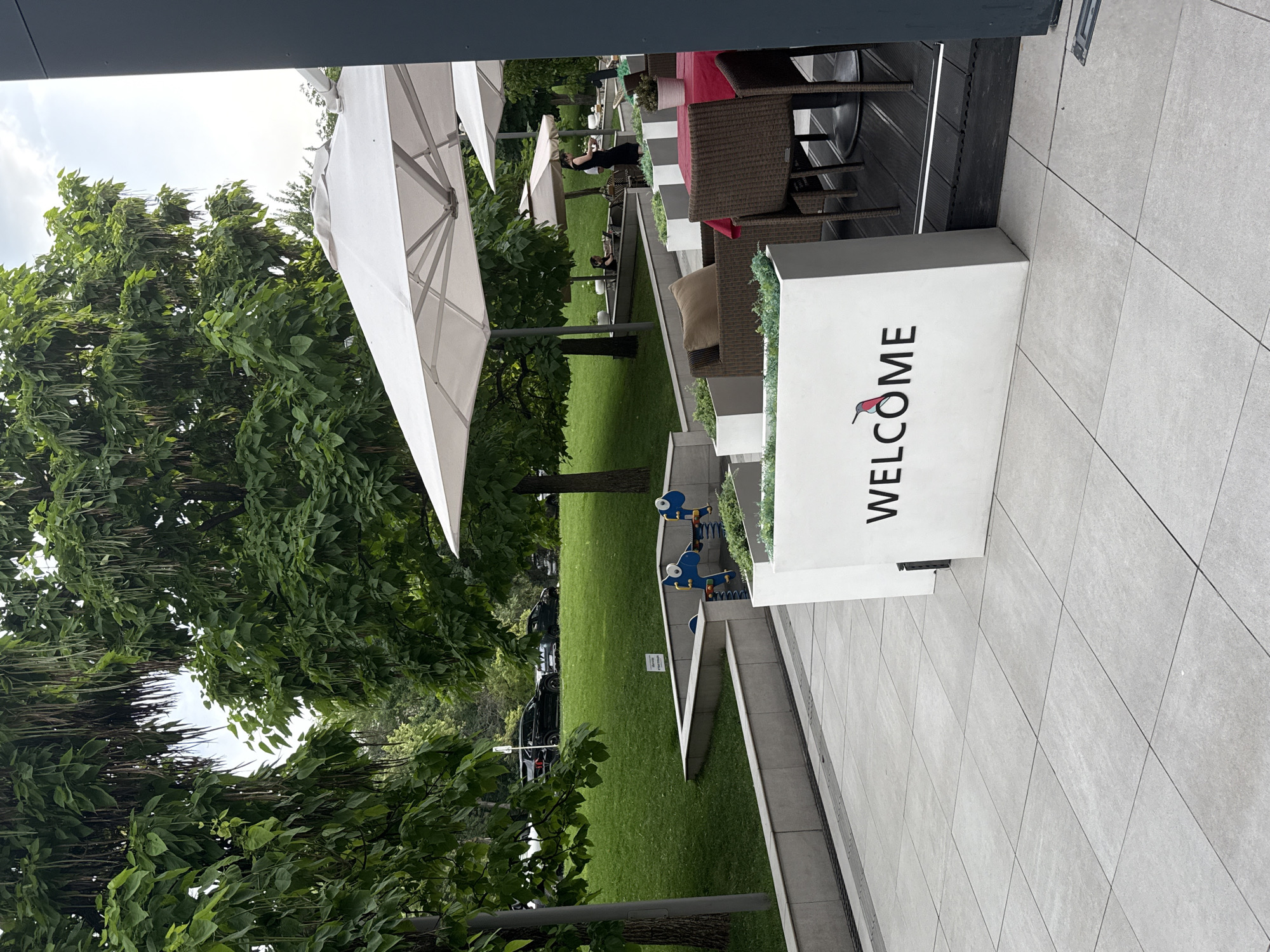 The image depicts an outdoor seating area of a cafe or restaurant in Almaty, Kazakhstan, likely during daylight hours. In the foreground, a light gray tiled patio features a white planter box with the word "WELCOME" and a red and black hummingbird logo. Beyond the planter, wicker chairs and tables are set up, one with a red tablecloth. Large white umbrellas provide shade. In the middle ground, a well-maintained green lawn slopes upwards, dotted with trees. A children's playground with blue spring riders is visible on the lawn. Cars are parked in the background. The overall environment suggests a pleasant, possibly upscale, outdoor recreational space.