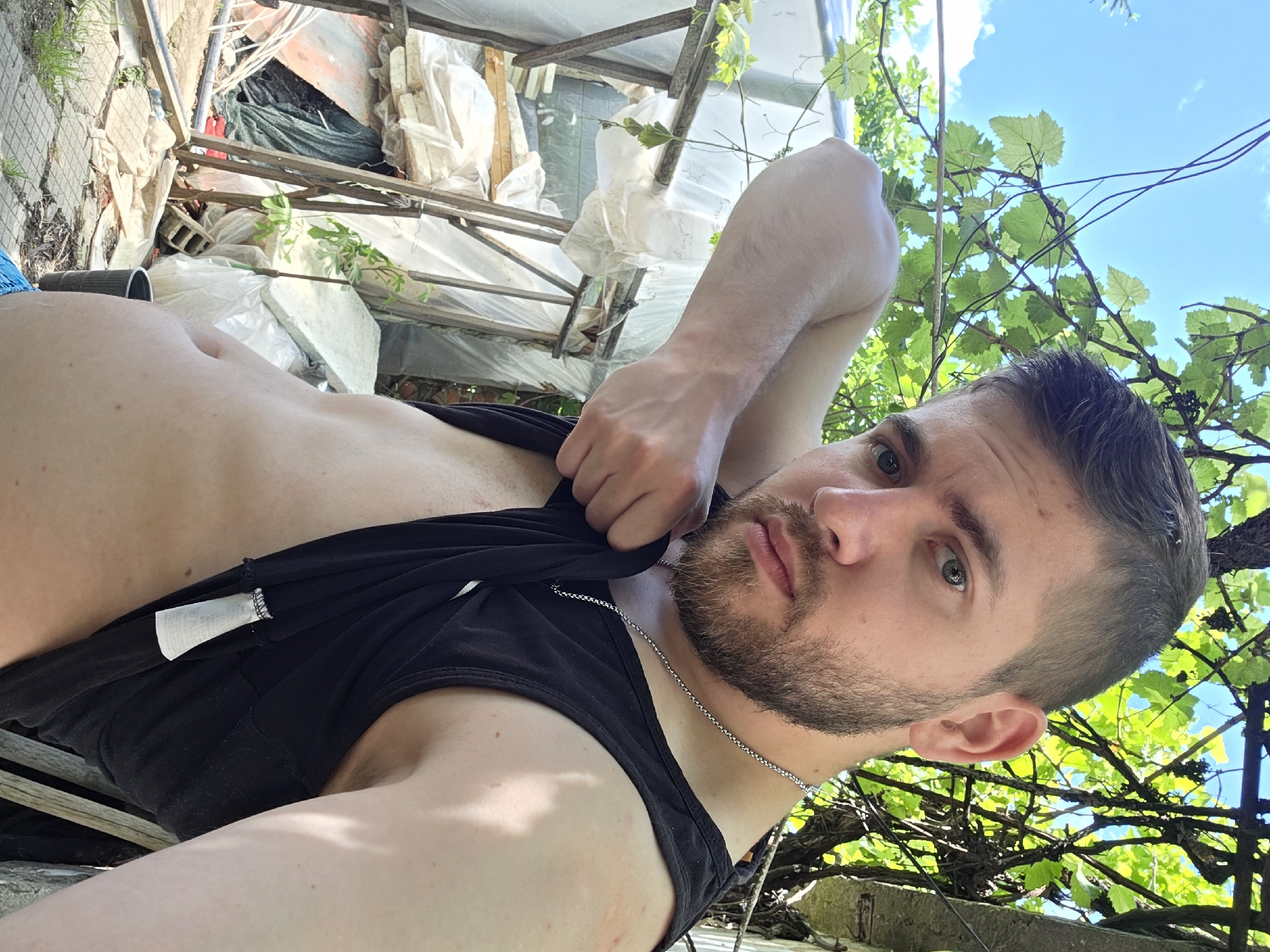 A man with short, dark hair, a beard, and a silver chain around his neck is holding up the bottom of his black tank top to reveal his torso. He appears to be in an outdoor area. Behind him is a structure constructed from white and metal materials, possibly a partial enclosure. Foliage, including grape vines, is visible against a background of blue sky. The location is in a yard near Yagodovo, Bulgaria.