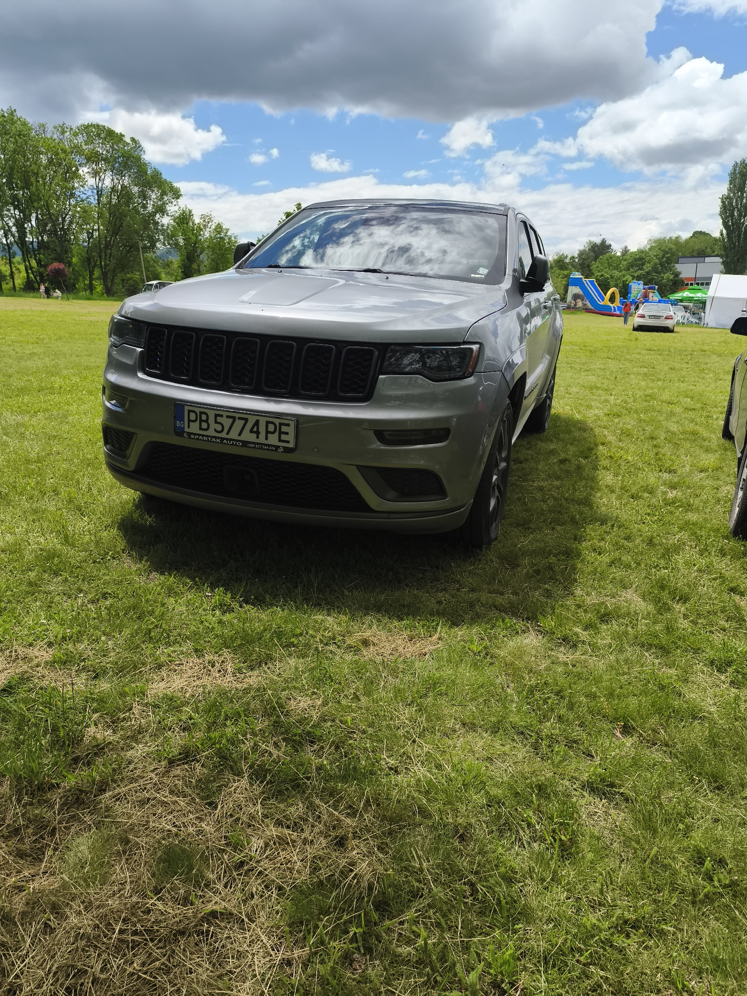 The image shows a silver Jeep SUV parked on a grassy field, likely in a park or recreational area. The car has a Bulgarian license plate reading "PB 5774 PE". In the background, there are trees, a blue and yellow inflatable slide, and a white car, which indicate the scene might be a public event or gathering. The sky is partly cloudy. There is no direct evidence to confirm that the location is Targovishte, Bulgaria, but the license plate suggests the vehicle is registered there.