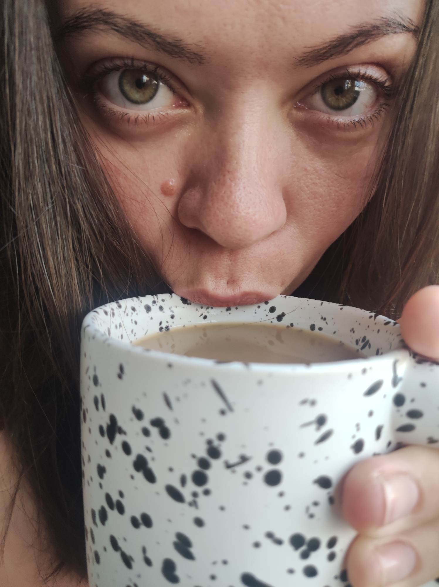 A close-up shot shows a woman with brown hair and hazel eyes drinking from a white mug with black splatter-like designs. The mug contains a light-colored beverage, likely coffee with milk. The woman's face is the primary focus, with her nose and pursed lips near the rim of the mug. The scene appears to be indoors, with no indication of the city of Chaikovsky, Russia.