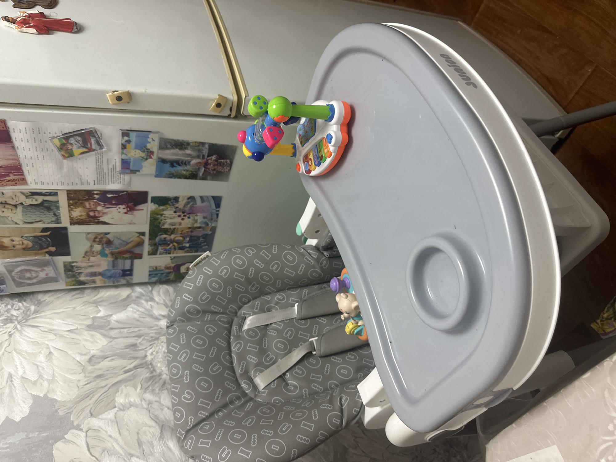 A high-angle medium shot captures a baby's high chair in a room, likely a home in Загородный, Russia, given the context. The high chair, upholstered in gray fabric with a repeating pattern of white circles and lines, features adjustable straps. A gray tray with a circular indentation for a cup is attached to the chair. On the tray rests a colorful baby toy, a musical keyboard with various buttons and a spinning element. In the background, a white refrigerator is adorned with numerous photographs and magnets. To the left of the high chair, a wall is covered with light-colored floral wallpaper. The floor beneath the chair appears to be made of wood.