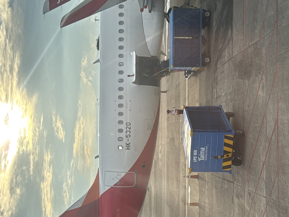The image showcases an airplane being serviced at an airport. The plane, marked with "HK-5320," has its rear cargo door open. Next to the aircraft are two blue cargo carts, one with diagonal yellow and black stripes. A person wearing a red and white reflective vest stands nearby. The backdrop features a bright, sunlit sky with scattered clouds. The photo is taken from an elevated perspective, possibly from within another aircraft.