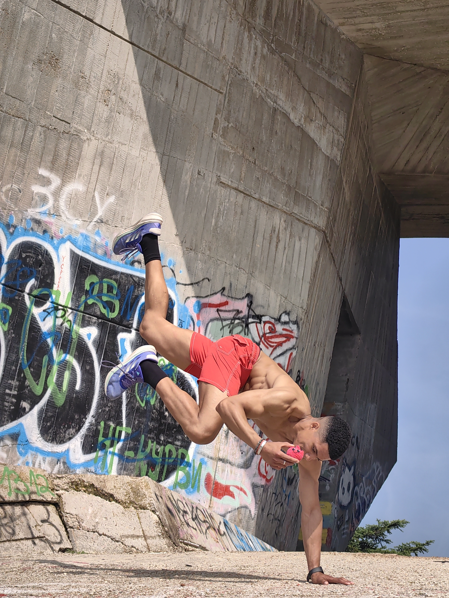 The image depicts a muscular, dark-skinned male with short, curly hair performing a one-armed handstand on a textured concrete surface. He is shirtless, wearing red athletic shorts, black socks, and blue and white athletic shoes. A black wristband is on his left wrist, and multiple bracelets are on his right wrist, which also bears a silver ring. His left hand is planted on the ground, while his body is inverted, with his legs bent at the knees and extending upwards and to the left. With his bent right arm, he holds a small, pink, spherical object with a smiling facial expression printed on it, bringing it close to his face as if inspecting or smelling it. He is looking downwards at this object.
The background features a massive, angular concrete structure, possibly part of a bridge or overpass, with visible formwork lines and textured surfaces. Extensive graffiti in various colors (blue, green, black, white, red) covers the concrete walls behind and to the left of the individual. A sharp diagonal shadow bisects the upper part of the concrete structure. In the upper-right corner, a clear blue sky is visible, along with a small section of green foliage from a coniferous tree. The ground is a light-colored, pebbled concrete surface. The scene is set in Varna, Bulgaria, under bright daylight conditions.