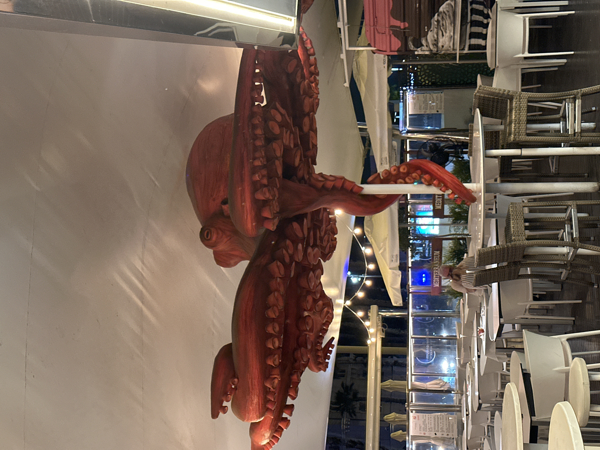 An overhead perspective captures a large, bright red, artificial octopus sculpture suspended from a white, tent-like canopy, dominating the upper half of the frame. Its numerous suction-cupped tentacles extend downwards and outwards. Below the octopus, an outdoor seating area for a restaurant or cafe is visible. Numerous white-topped tables and white chairs are arranged neatly. A few bar stools with woven seats are also present. In the background, glass-paneled walls with visible signage can be seen, including a sign that reads "MEDITERRANEUM". String lights hang across the canopy. One person, seen from the back, is seated at a table in the middle ground, wearing a striped top. The overall lighting suggests it is evening or dusk.