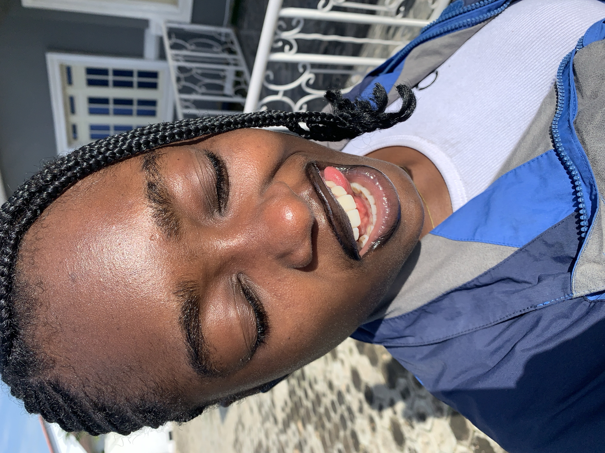 A young Black woman with braided hair and closed eyes playfully sticks out her tongue, her lips glossy and dark. She is wearing a blue and grey jacket zipped over a white ribbed tank top. The background shows a white railing and a building with blue-paned windows, suggesting an outdoor setting in a developed area, possibly a residential building in Gura Top, Nigeria. The ground beneath her is paved and textured, with shadows cast by sunlight.
