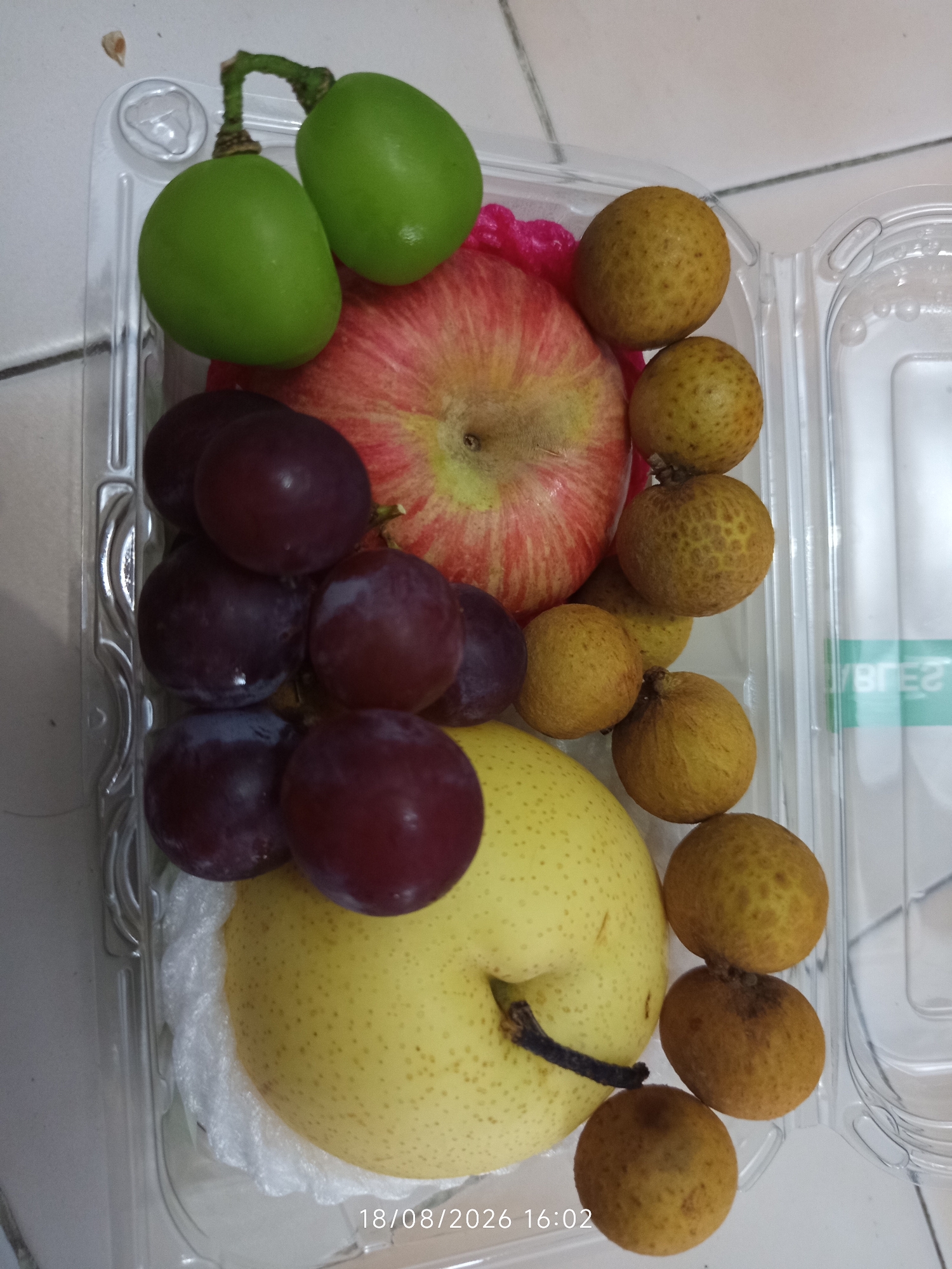 A clear plastic container holds a fruit assortment. The fruits include green grapes, red grapes, a red apple, a yellow pear, and several small, round, tan-colored fruits that appear to be longans. The container is filled with these fruits, with some overlap. The background consists of a light-colored tiled surface. A timestamp "18/08/2026 16:02" is visible in the lower right corner of the image. The scene appears to be an indoor setting.