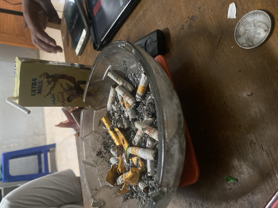 The image captures an indoor scene in the city of Cimenyan, Indonesia, focusing on a cluttered wooden table. A clear, rectangular glass ashtray, heavily laden with dark grey ash, numerous white-filtered cigarette butts, and crumpled gold foil, occupies the center foreground. This ashtray rests on an orange, possibly plastic, base. To its immediate right stands a yellow Ultra Milk carton, featuring the text "Make Your Move!" and an illustration of two figures, with a white, bent drinking straw inserted.
Further right on the table, three mobile devices are present: a black tablet, a black smartphone, and a white smartphone. A black remote control lies beside the ashtray on the right. In the lower right corner of the table, a tarnished silver-colored coin and a small, torn piece of white paper are visible. A small crumpled piece of green material is located in the lower left corner.
The upper left portion of the frame shows the arm and torso of an individual wearing a white shirt, seated at the table. On the right side, the right hand of another individual is visible, positioned near the mobile devices. The background features a light-colored wall, with a blue plastic stool visible in the upper left and part of a wooden door or furniture item in the upper right. The floor, partially visible, appears to be tiled.