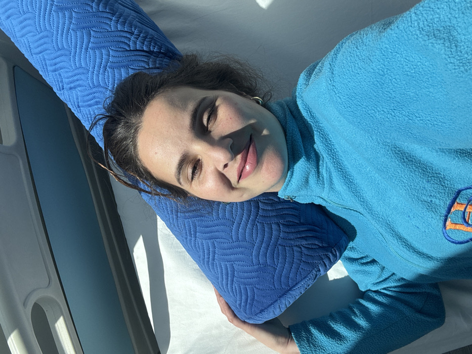 The image displays a female subject, appearing to be in her 20s or 30s, lying down and facing the camera. She has dark brown hair, brown eyes, and is smiling with slightly parted lips adorned with light pink lip gloss. Small silver hoop earrings are visible on both ears. The woman is wearing a bright turquoise fleece quarter-zip pullover, which features an orange and white embroidered "H" logo on the left chest area. Her left arm (viewer's right) holds a blue cylindrical bolster pillow with a distinct quilted wave pattern, positioned behind her head and neck. She is resting on a white sheet or light-colored fabric. The background reveals the interior of what appears to be a vehicle, characterized by a light grey overhead panel with a curved handle-like fixture and a horizontal window showing a light blue tint. The scene is illuminated by direct sunlight, casting noticeable shadows on the white fabric and the woman's face. The image contains no discernible visual information to indicate the city of Meknes, Morocco, or any other specific geographical location.