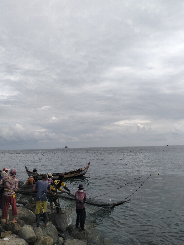 A group of people are fishing near a rocky shoreline. They are pulling a large net from the water, presumably filled with their catch. A small, colorful boat rests nearby. The sea is calm, with a few distant vessels visible on the horizon. The sky is overcast with gray clouds, suggesting a potentially stormy weather. The scene captures a moment of traditional fishing activity in a coastal environment.