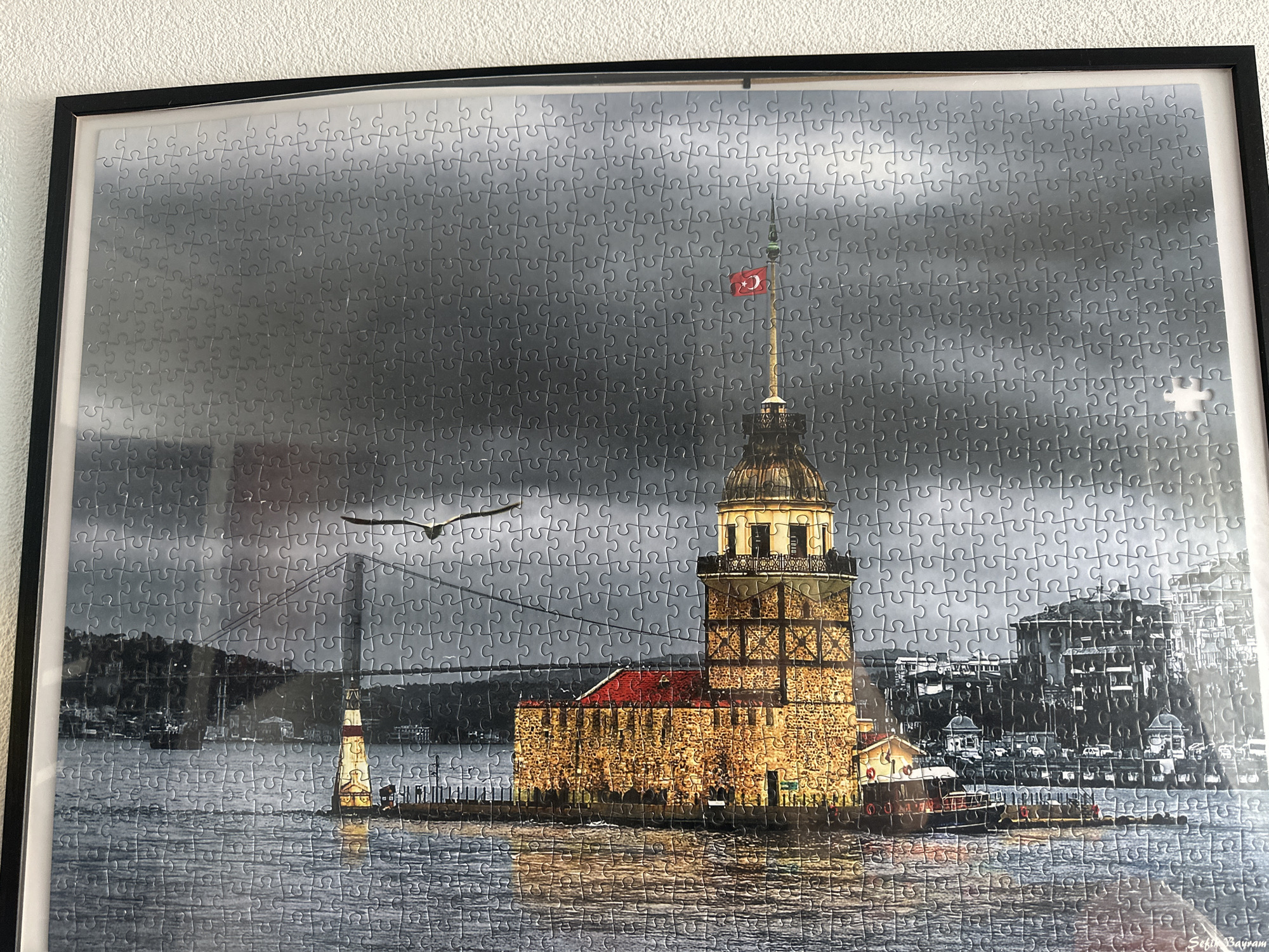 The image is a framed jigsaw puzzle depicting a nighttime scene of the Maiden's Tower (Kız Kulesi) in Istanbul, Turkey, illuminated and reflected in the water. The tower is centrally located, with a Turkish flag flying from its spire. To the left, a bridge and a lighthouse are visible across the water, and a bird is flying in the sky. On the right, the silhouette of a city skyline is present. The sky is overcast and dark, with the water showing ripples and reflections of the lights. The overall image is rendered in grayscale with the exception of the red roof of the tower and the Turkish flag. The puzzle itself is slightly askew within its black frame, which is hanging on a textured white wall. There are no people visible in the image. The image does not depict Günzburg, Germany.