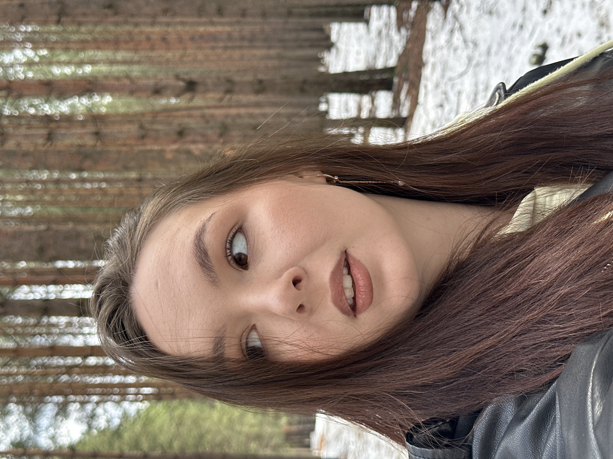 A young woman with long, dark brown hair poses for a selfie. She's wearing a dark leather jacket. The background shows a forest scene with tall trees. Traces of snow can be seen on the ground between the trees. The location is likely a wooded area, possibly a park or forest, in or near the city of Chaikovsky, Russia.