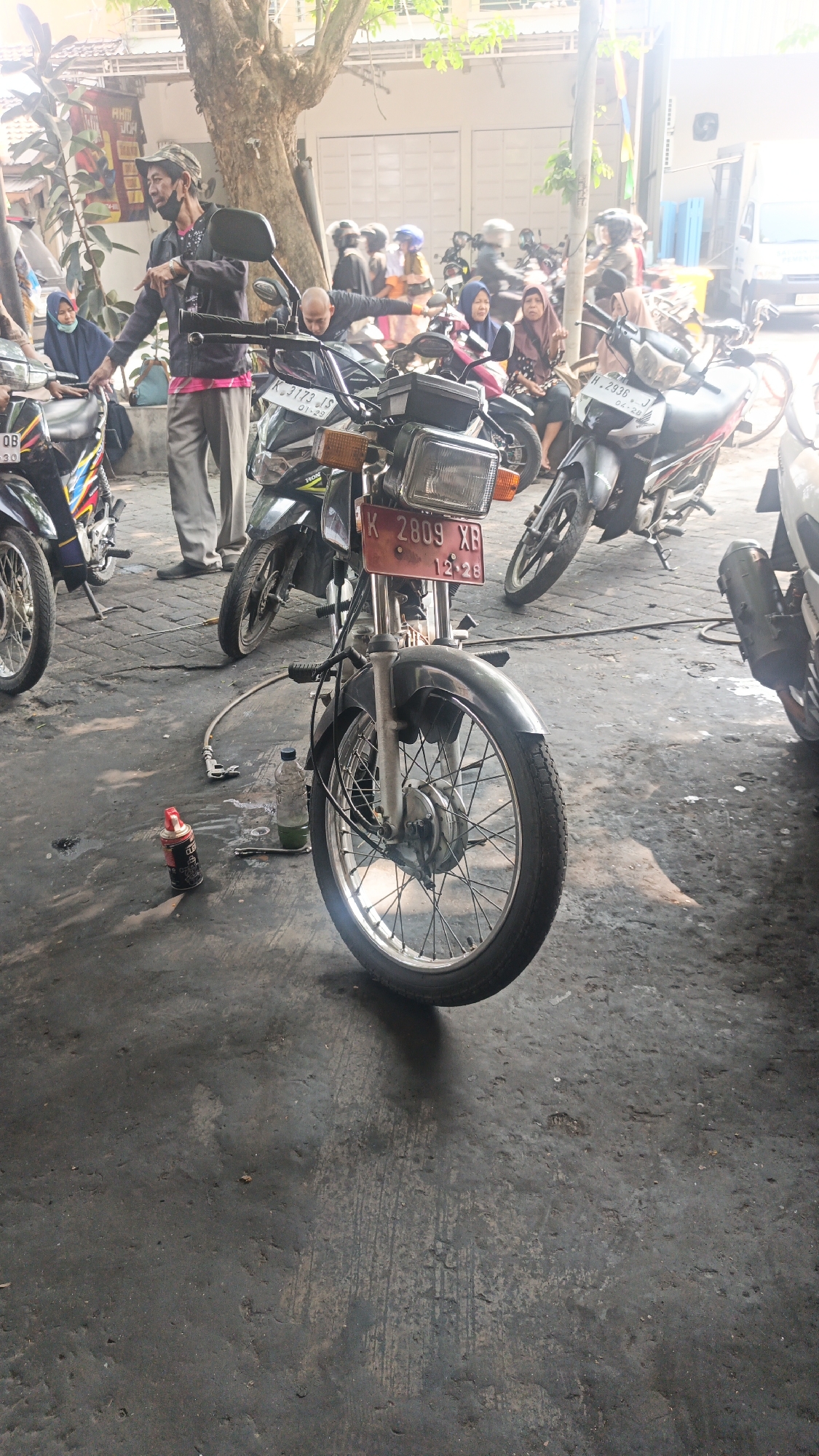 The image depicts an outdoor scene in what appears to be a motorcycle repair area or parking lot in Mlati Kidul, Indonesia. A man wearing a cap and a dark jacket over a pink shirt, along with gray trousers, is standing and gesturing with his left hand. Several motorcycles are parked around the area. In the foreground, a motorcycle with a red license plate displaying "K 2809 XB 12.28" is prominently featured, its front wheel and fender clearly visible. On the ground near this motorcycle are a bottle of liquid, a can, and some tools, suggesting repair or maintenance activities. In the background, other individuals, some wearing headscarves and helmets, are visible near other motorcycles. The environment is a paved ground surface, likely concrete, with some debris and oil stains. A large tree is present on the left side of the image, and structures with white doors and a white van are visible in the background, indicating a commercial or residential area. The lighting suggests it is daytime, with sunlight filtering through the trees.