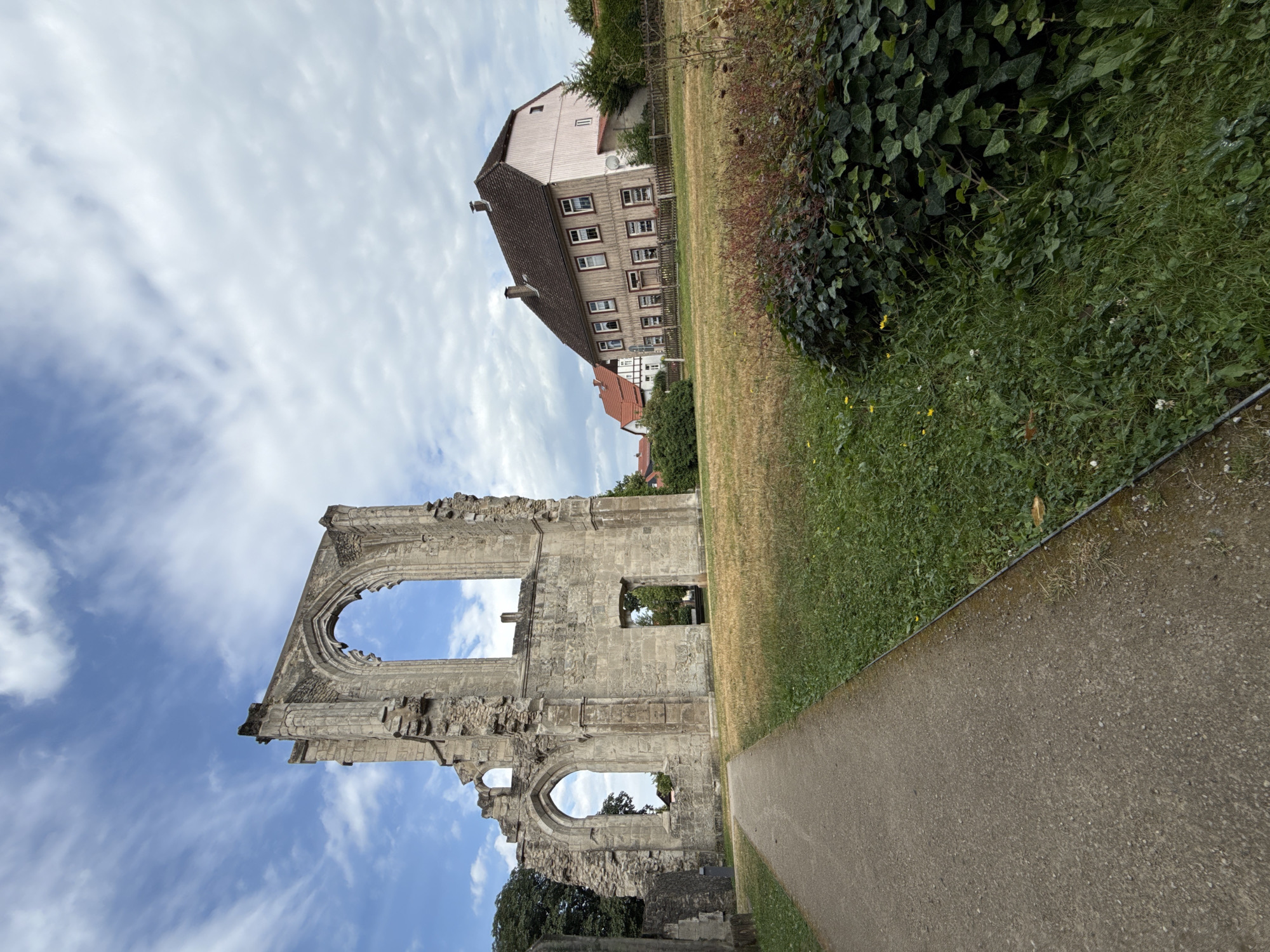 The image depicts the ruins of a Gothic-style building, possibly a former monastery or church, in Walkenried, Germany. The stone structure is partially collapsed, with large arched openings and remnants of intricate stonework visible against a cloudy sky. A paved path leads towards the ruins from the left foreground, bordered by a grassy area. In the background, several houses with tiled roofs are visible, suggesting a residential area adjacent to the historical site. Green trees and vegetation are scattered throughout the scene, some of which appear to be overgrown around the ruins and fences. The overall impression is one of historical preservation and integration with the modern town.