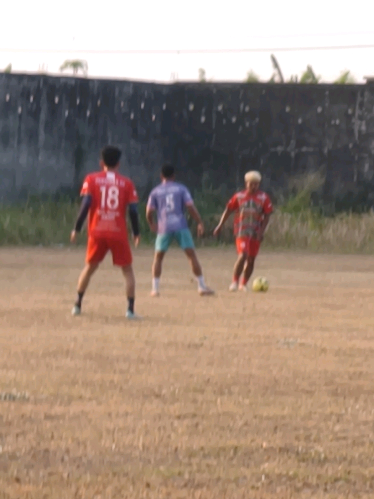 Three individuals are playing soccer on a dirt field in Desa Sindon, Indonesia. The field is dry and appears to be unmaintained, with patches of sparse grass and debris. In the background, a dark, textured wall runs horizontally, with a lighter sky above.
The person on the left is wearing a red jersey with the number "18" on the back and red shorts. They are facing away from the camera with their arms slightly bent.
The person in the middle is wearing a purple t-shirt and blue shorts. They are also facing away from the camera, with their arms behind their back.
The person on the right is wearing a red and white patterned jersey and red shorts. They are facing towards the camera and appear to be in the process of kicking a yellow soccer ball. Their hair is light-colored and styled upwards.
All three individuals are dressed in athletic attire. The lighting suggests it is daytime, possibly late afternoon due to the warm tones of the field.