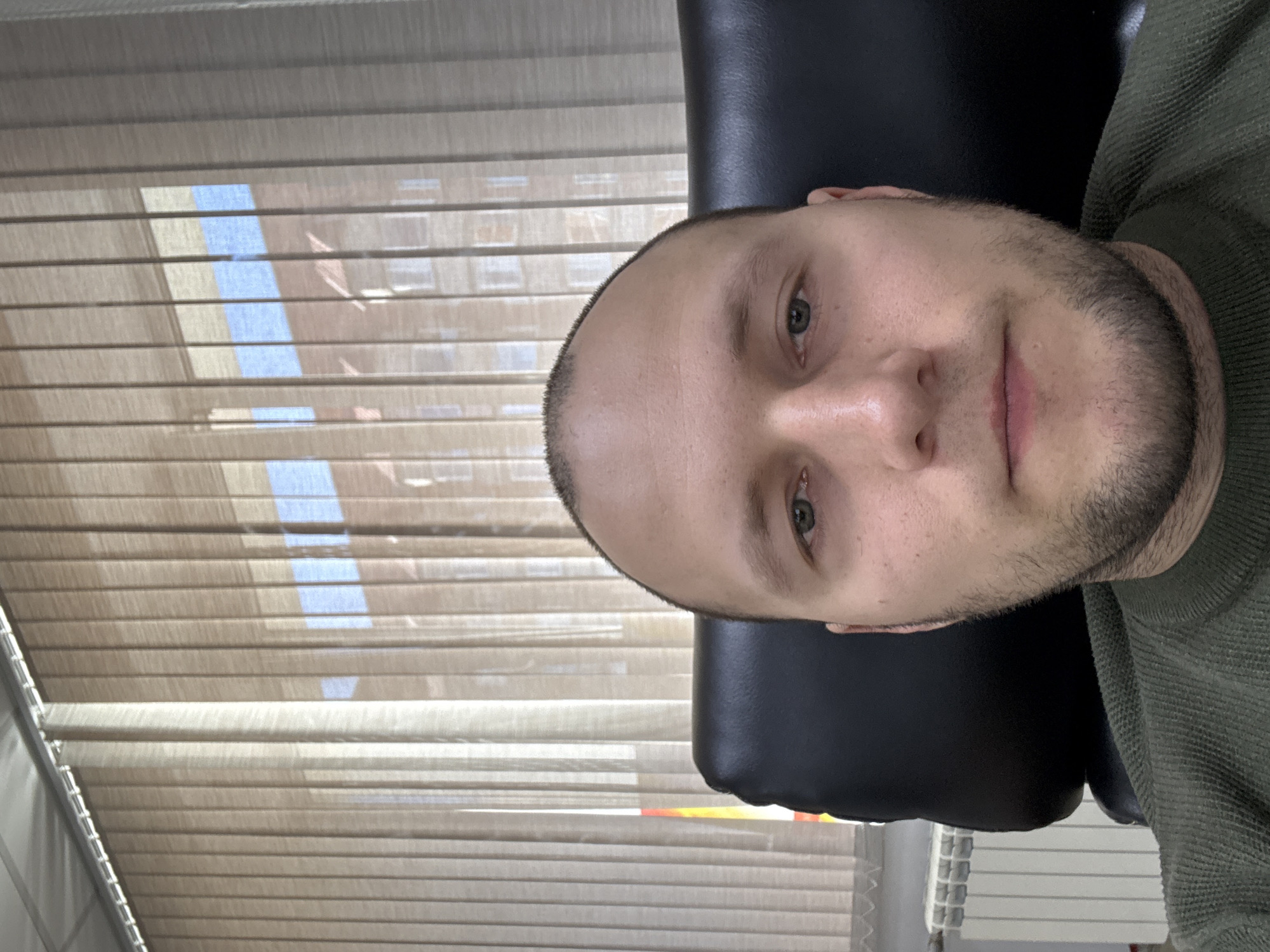A man is sitting in a black leather chair in front of a window with beige blinds. Light streams through the blinds, revealing a glimpse of blue sky. The man has short hair and a stubbly beard. He wears a green sweater. There is no information in the image to indicate the location in Krasnoyarsk, Russia.